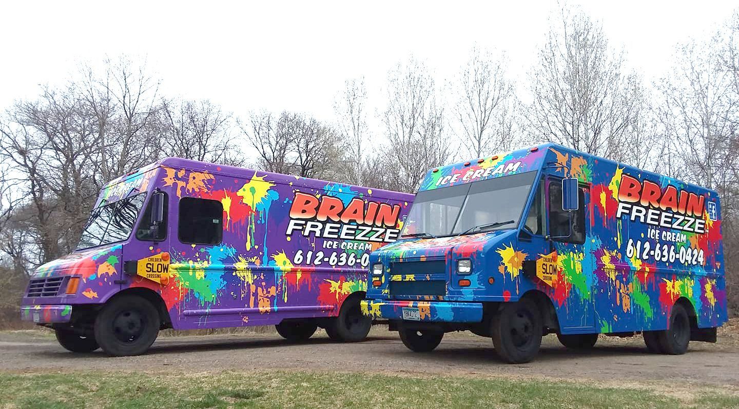 Two colorful freeze-dryer vans parked on grass in a wooded area