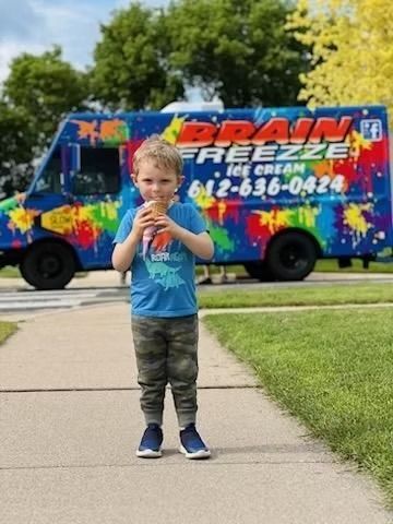 Child standing on a sidewalk in front of a colorful “Ice Breeze” truck, eating a treat.