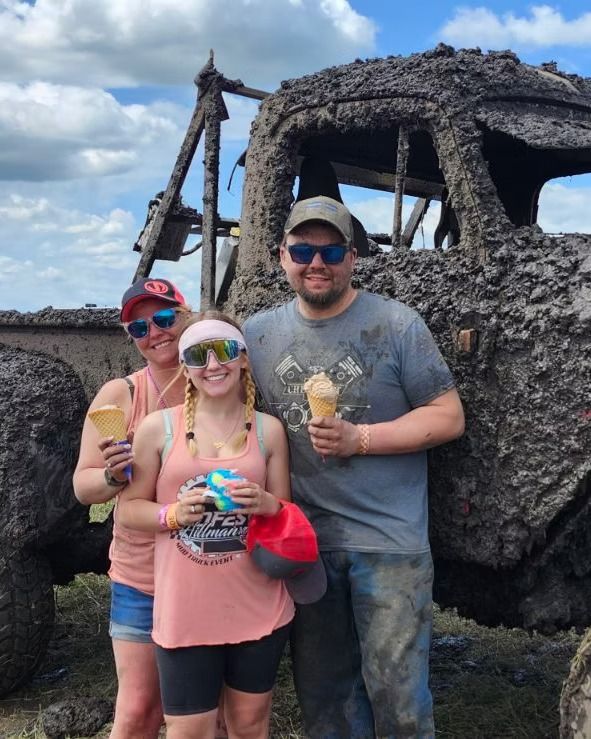 Three people posing with ice cream beside a weathered truck under a blue sky