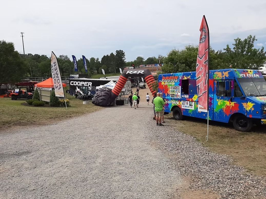 Gravel path at an outdoor fair with colorful food trucks and tall flags under a cloudy sky