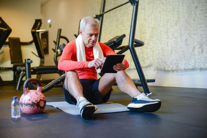 An elderly man is sitting on the floor in a gym using a tablet.