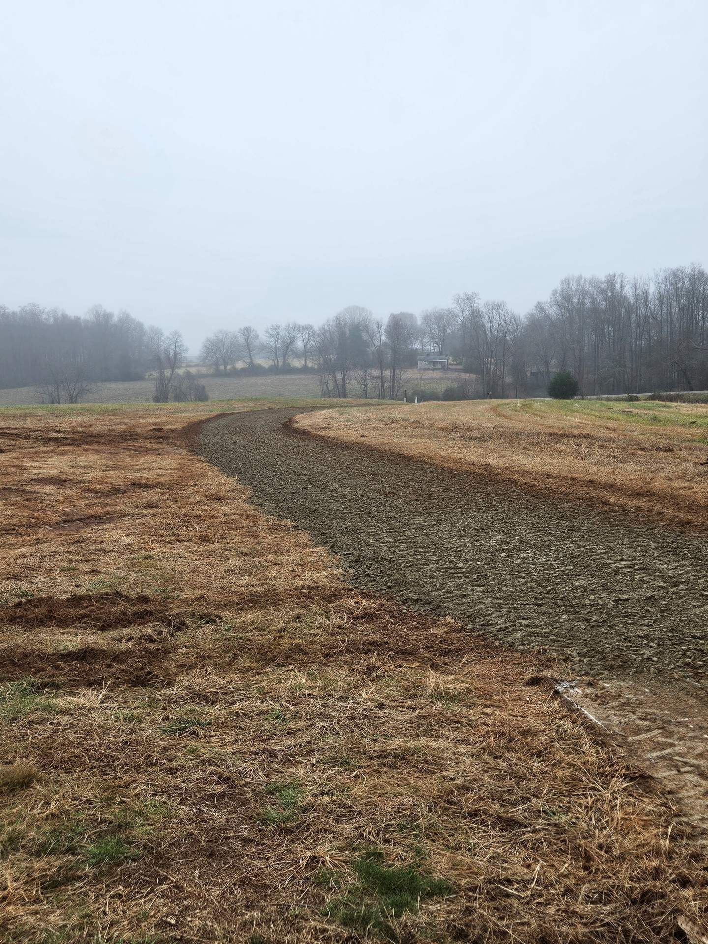 Bare dirt path winding through brown fields under a gray, overcast sky, with leafless trees in the distance