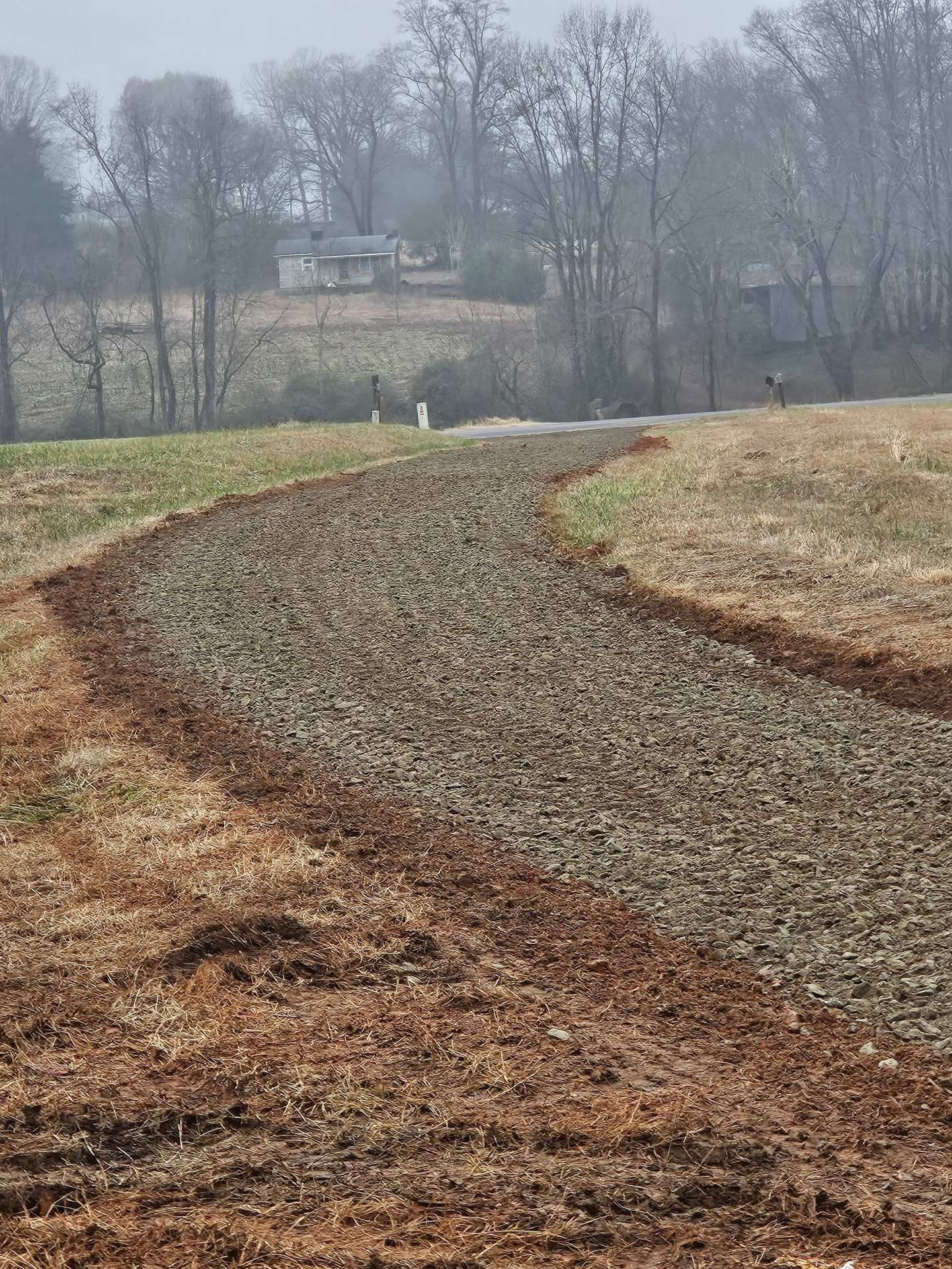 Curving dirt path through a brown field toward distant trees under a gray, foggy sky