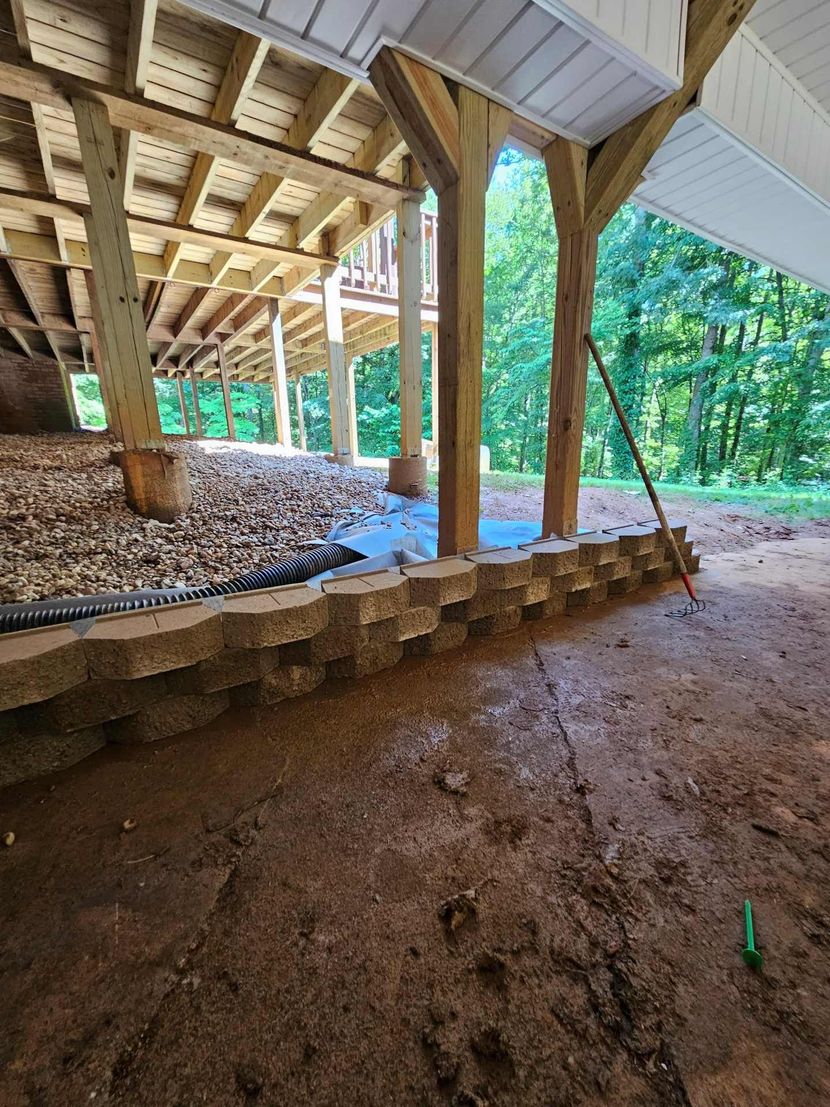 Wooden pavilion with posts and beams over a dirt floor, bordered by stacked stone edging and forest beyond