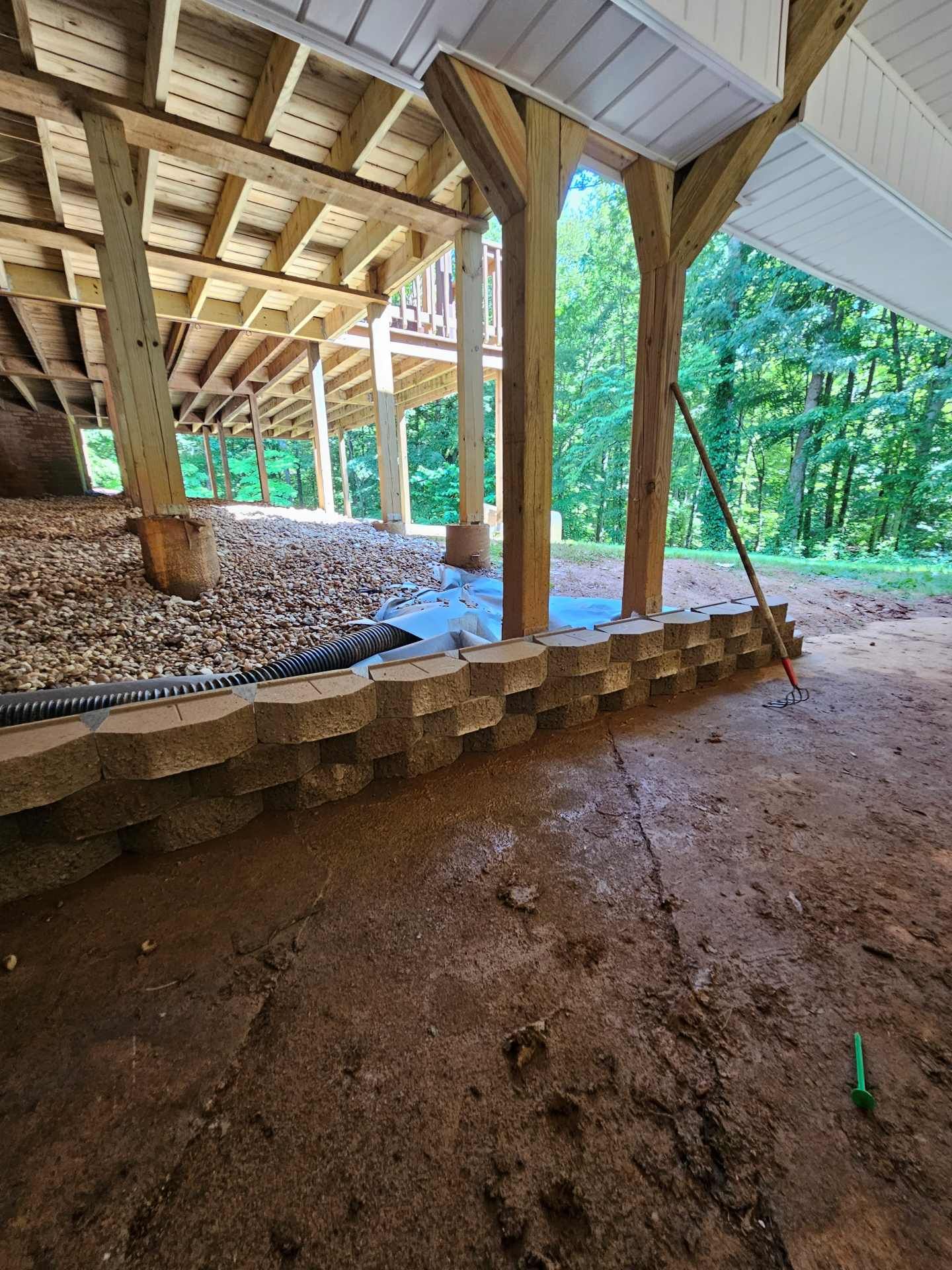 Under a raised pavilion, wooden posts stand over a gravel floor beside a forested area.