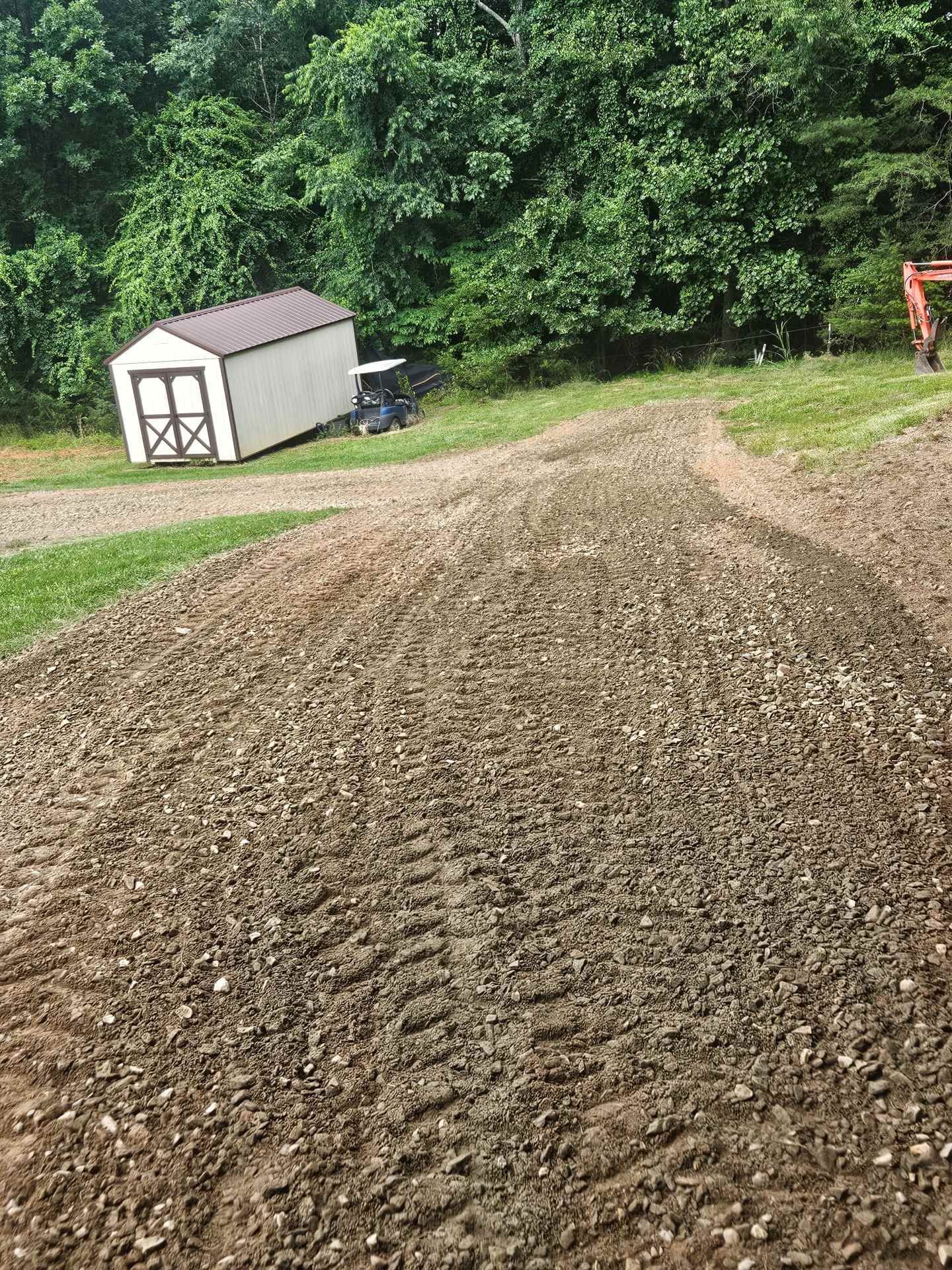 Gravel driveway beside a small white shed and dense green trees on a rural property.