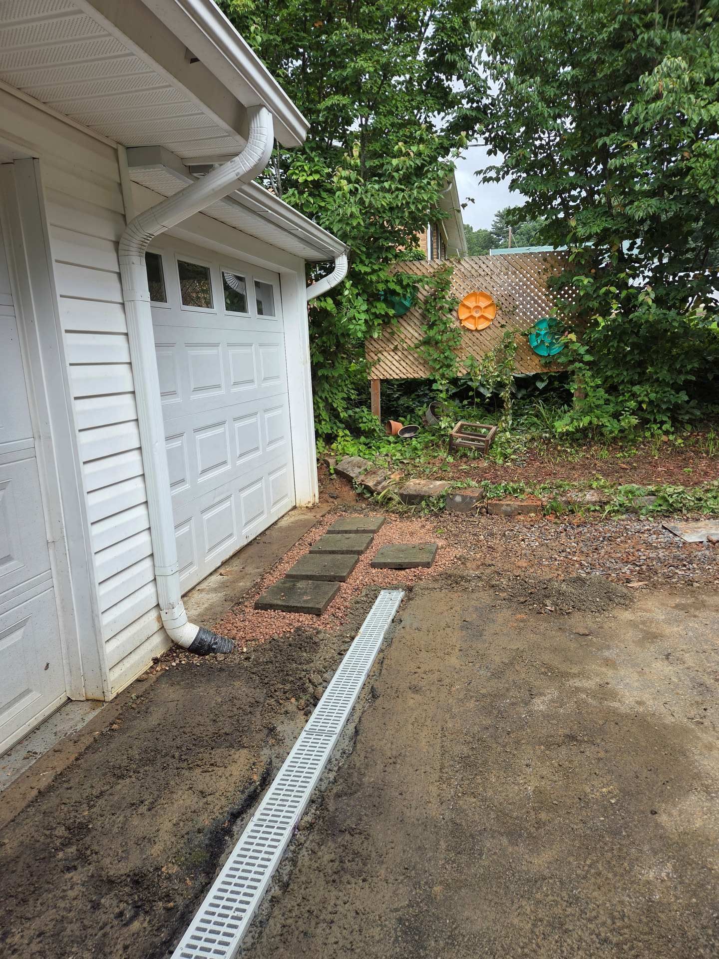 Side of white garage with driveway drain and wet concrete, backed by trees and a shed in the yard