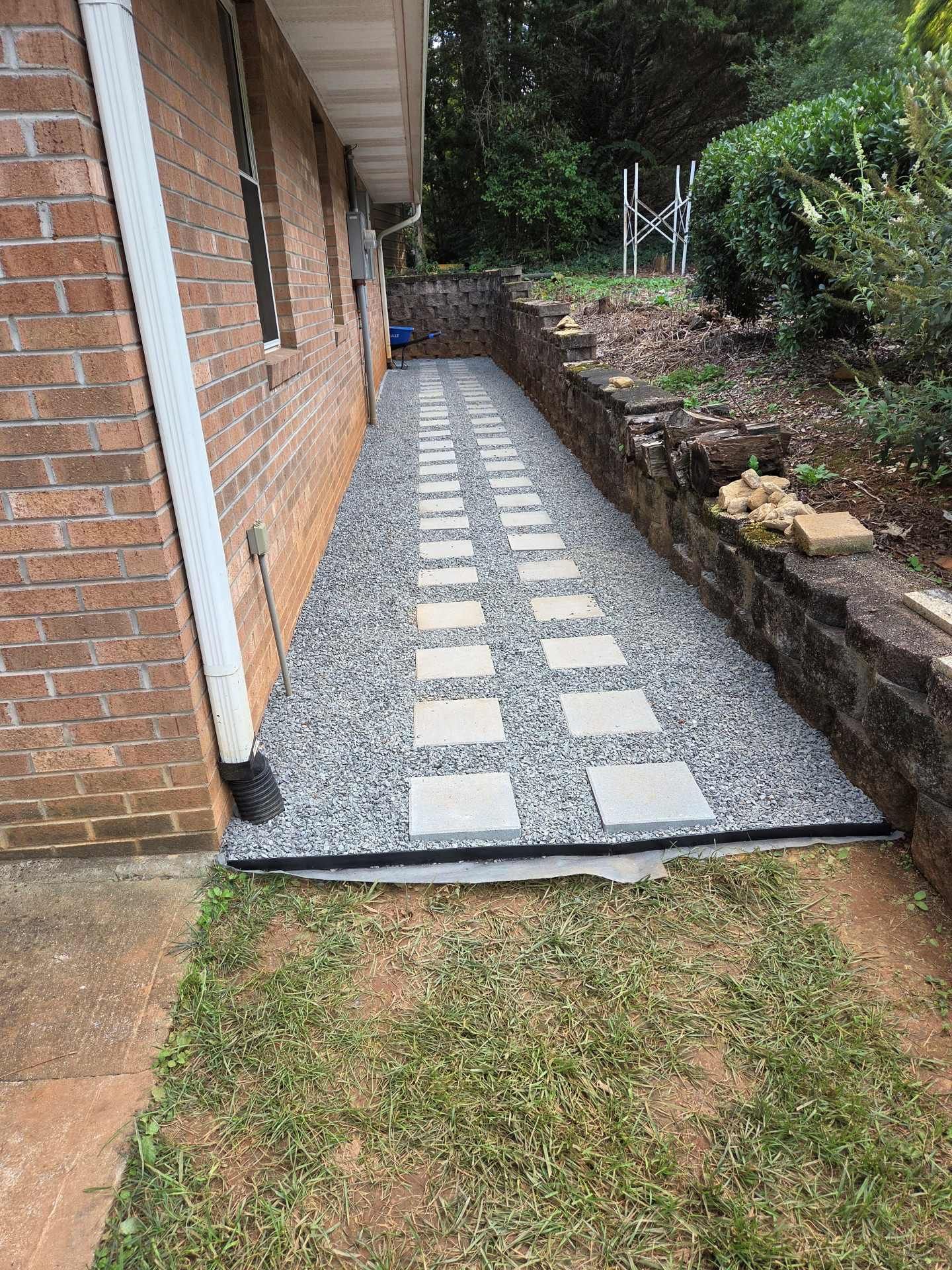 Narrow gravel walkway with stepping stones beside a brick house and stone retaining wall in a backyard.