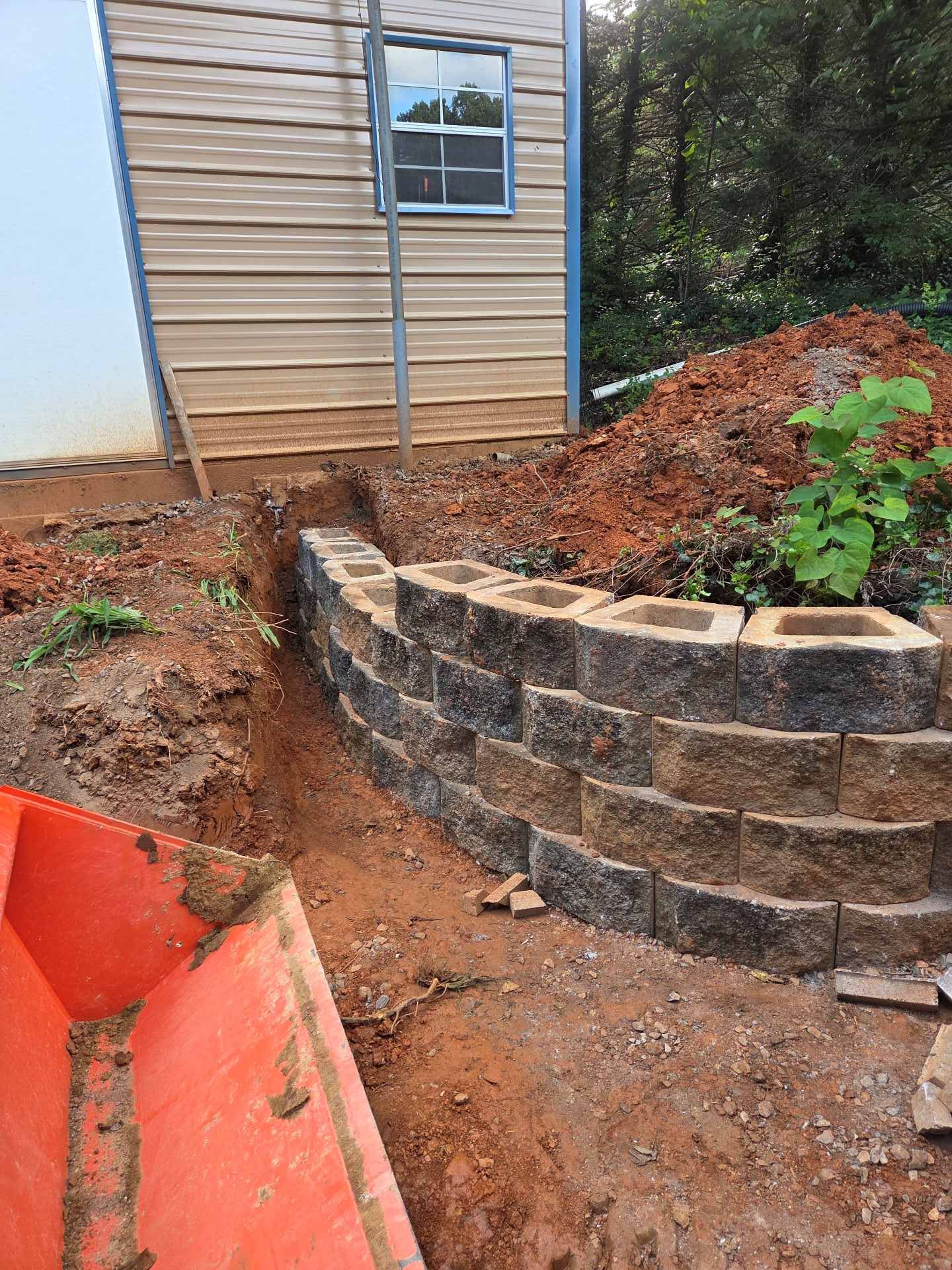 Backyard retaining wall under construction beside a tan shed, with red soil and a narrow dirt path