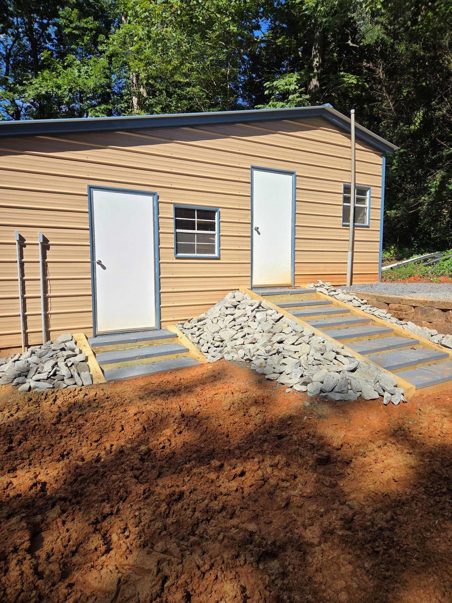 Small tan building with two white doors, a window, and gravel ramps under construction in a wooded area.