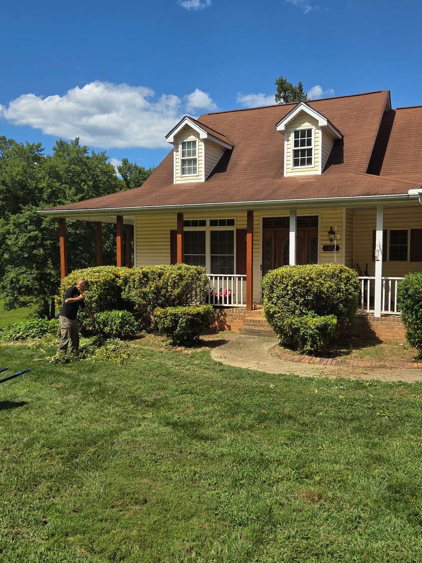 A tan house with a red roof and porch, with a person mowing the green lawn on a sunny day.