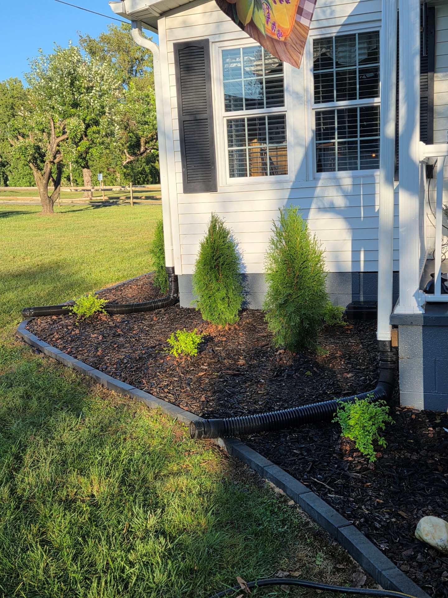 House corner with windows and a mulched flower bed bordered by black edging, beside a grassy yard.