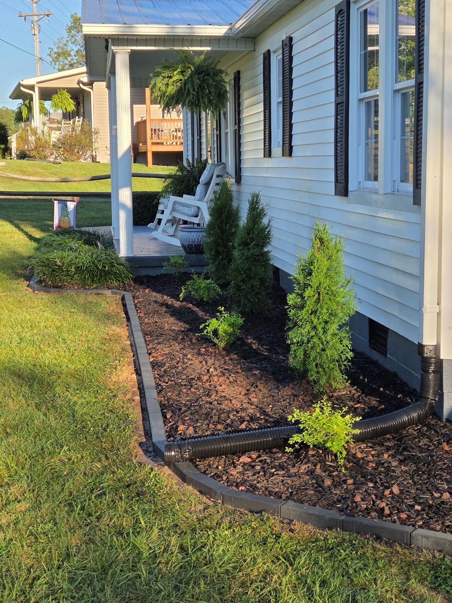 Front porch and side yard of a white house with black-trimmed windows, mulch beds, and small shrubs.