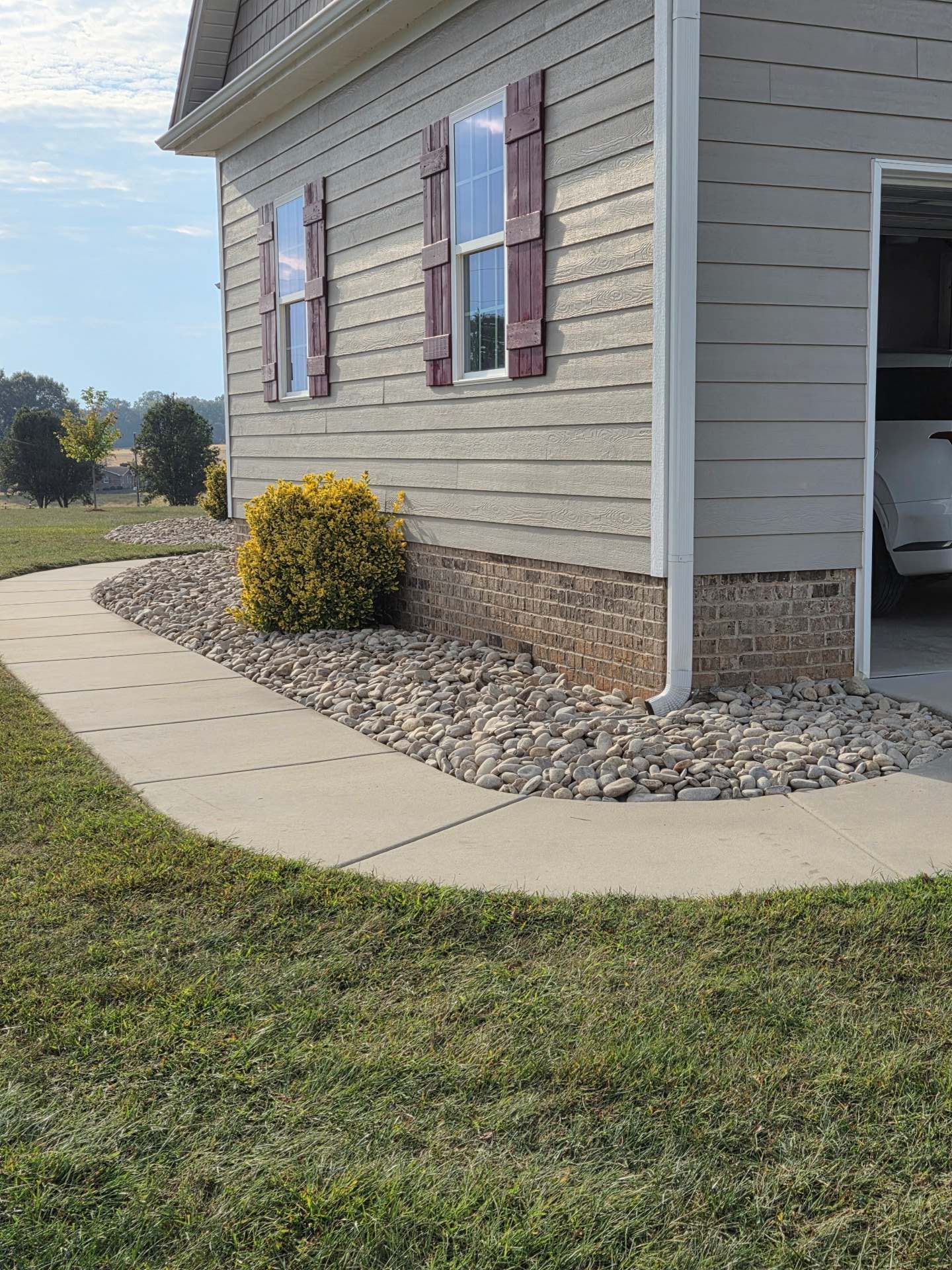 Suburban house corner with curved concrete sidewalk, rock landscaping, and beige siding with maroon shutters