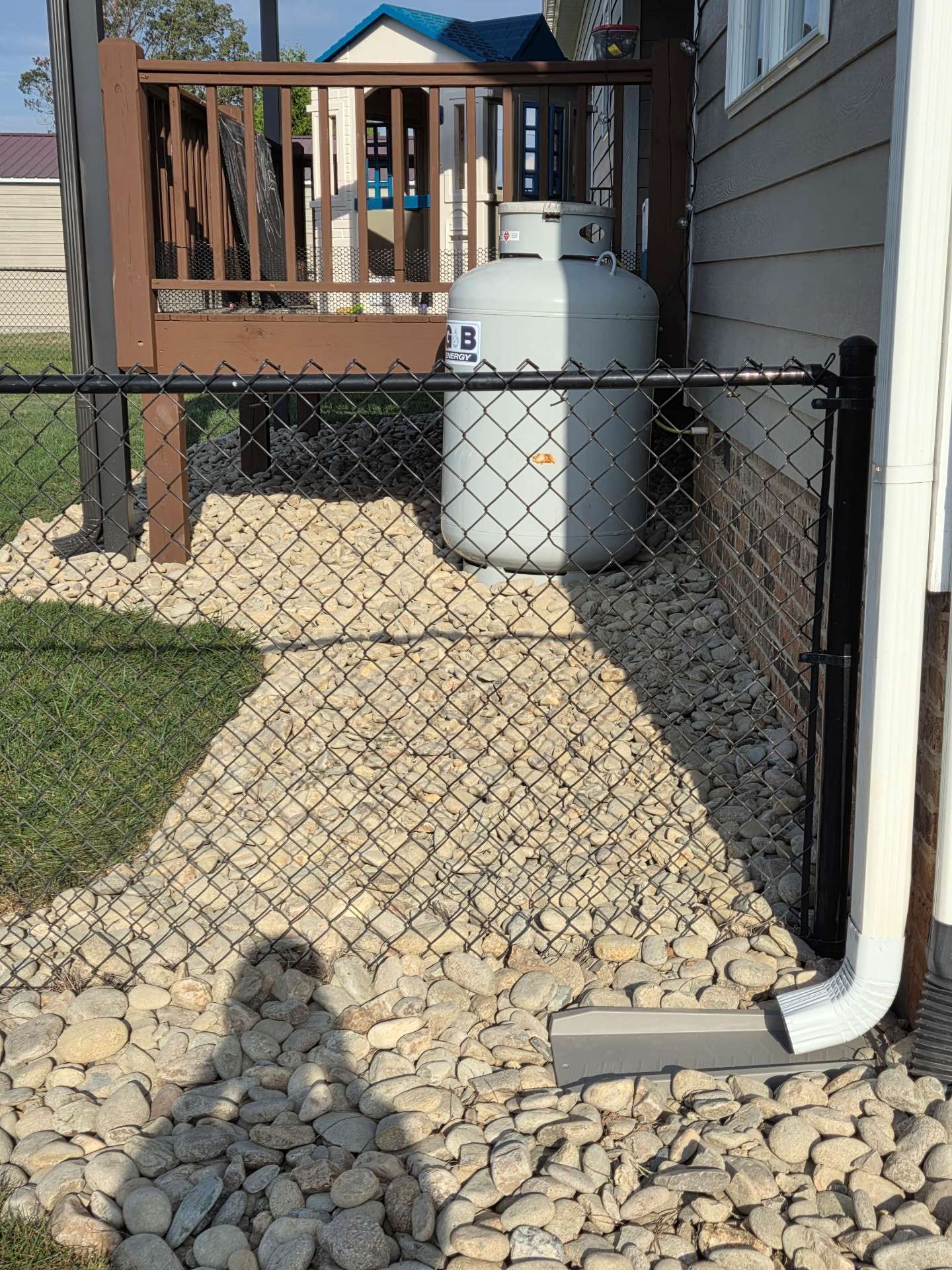 Chain-link fence gate beside a gravel walkway, with a propane tank and house porch in the background