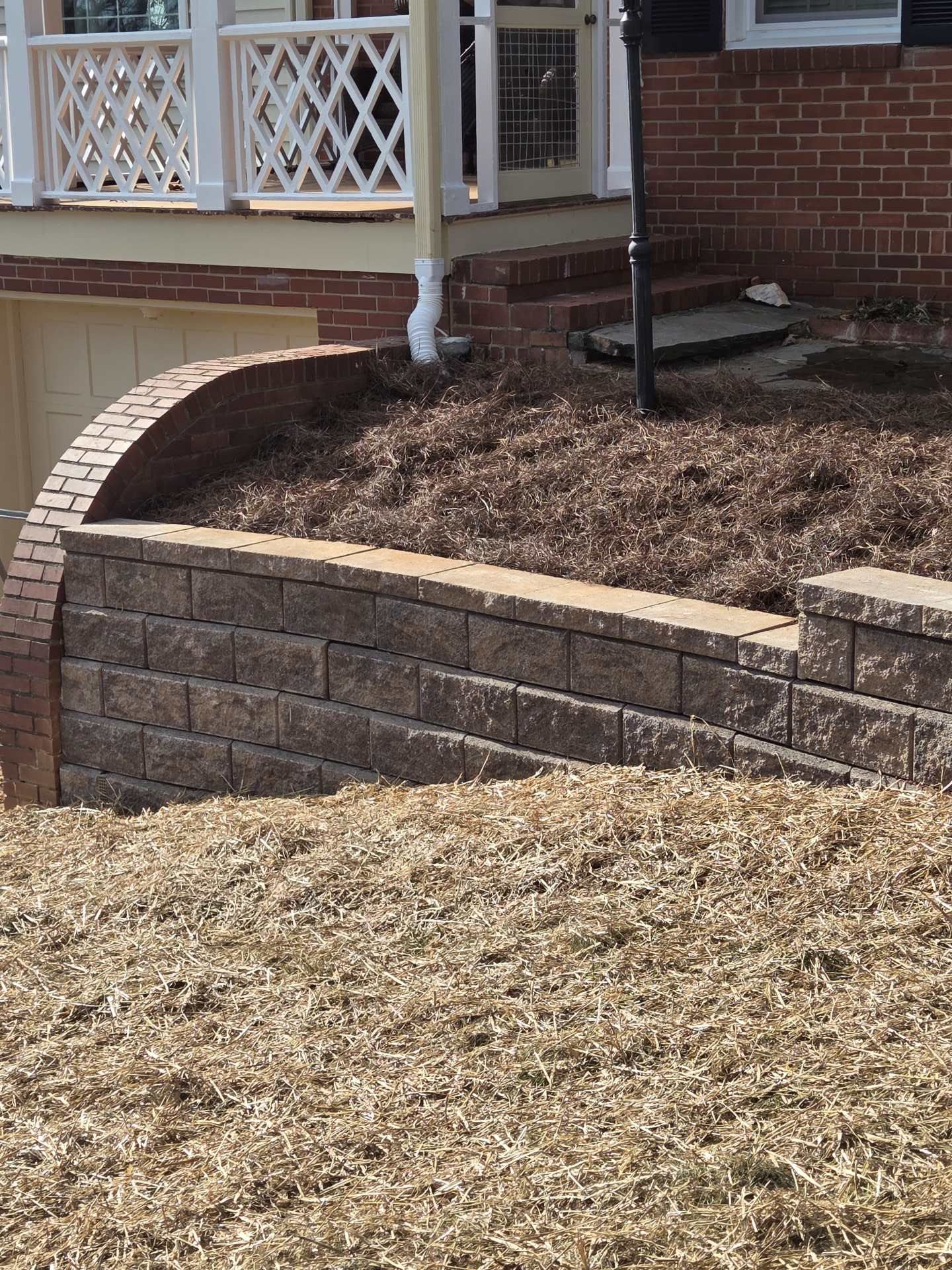 Brick retaining wall with mulched garden bed and porch railing beside a house