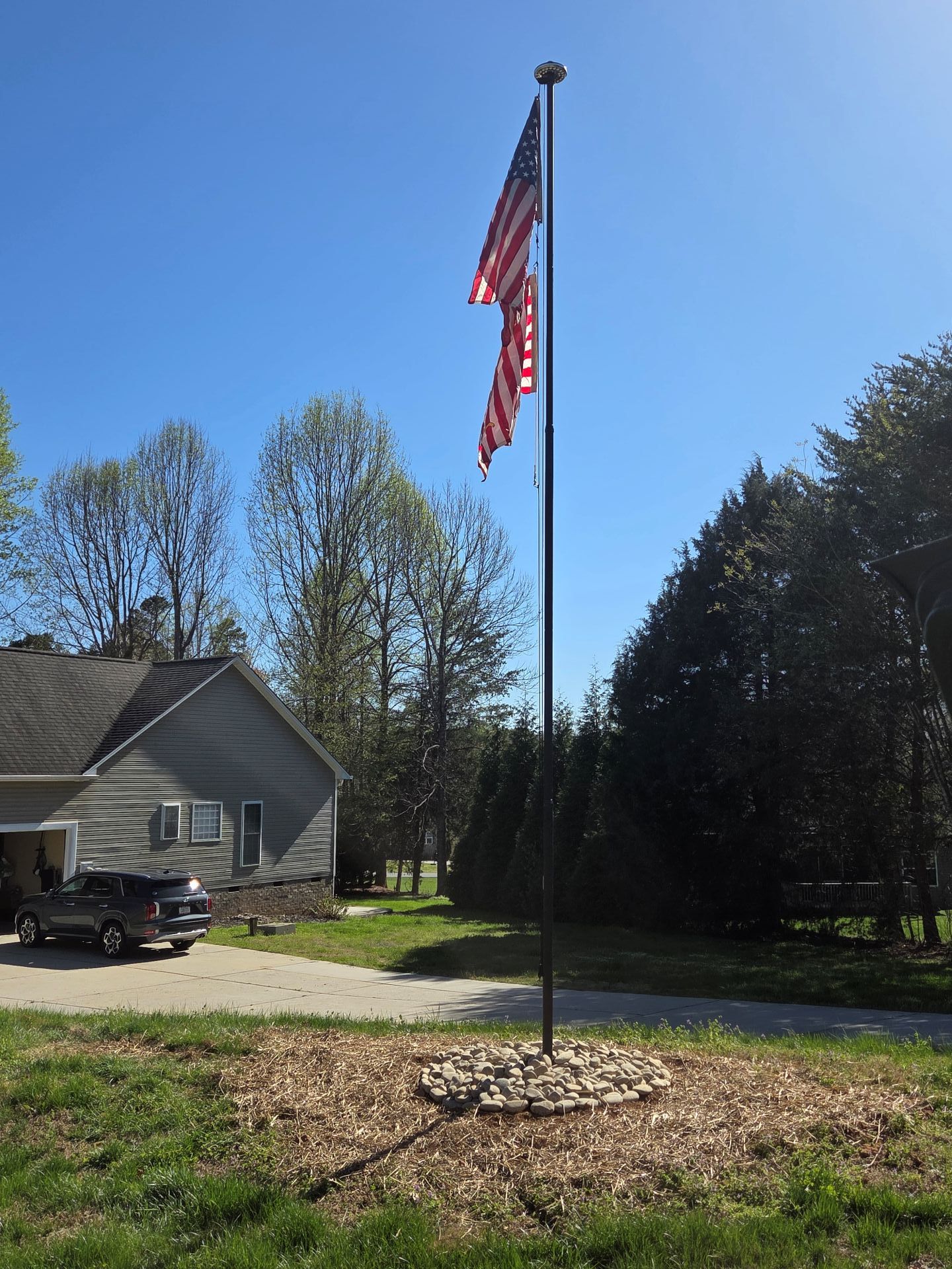 American flag on a tall pole in a yard, with a house, gravel driveway, and trees under a blue sky