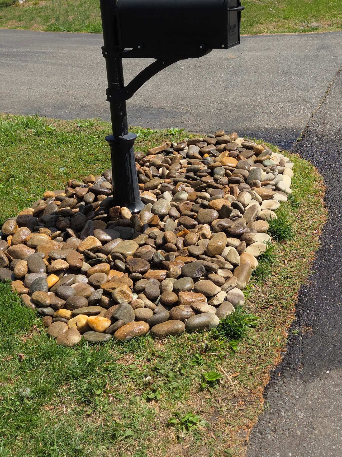 Black mailbox on a post beside a rock-covered border in a grassy yard by the road