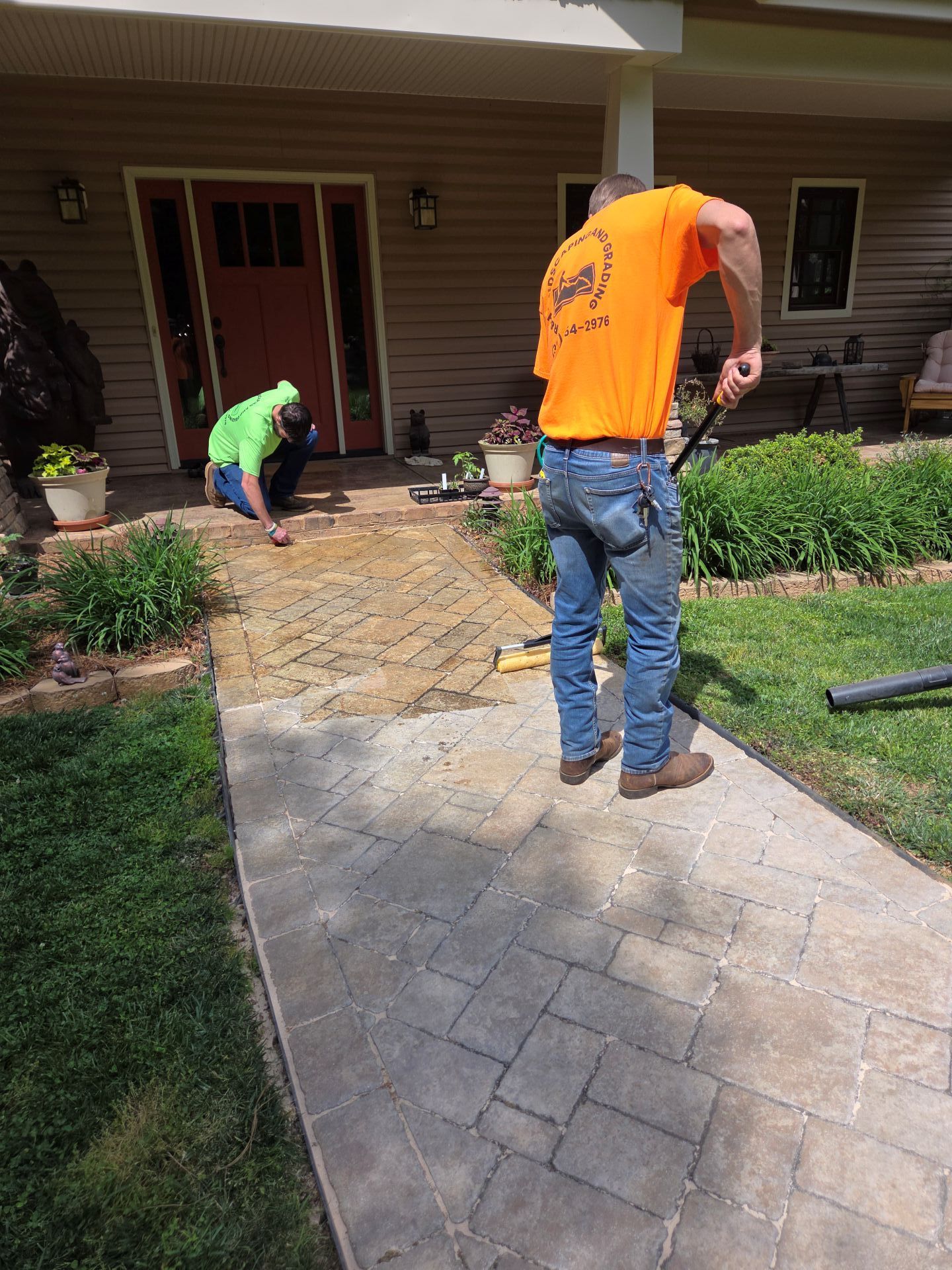 Two workers installing a stone walkway in front of a house, one in an orange vest and one kneeling in green.