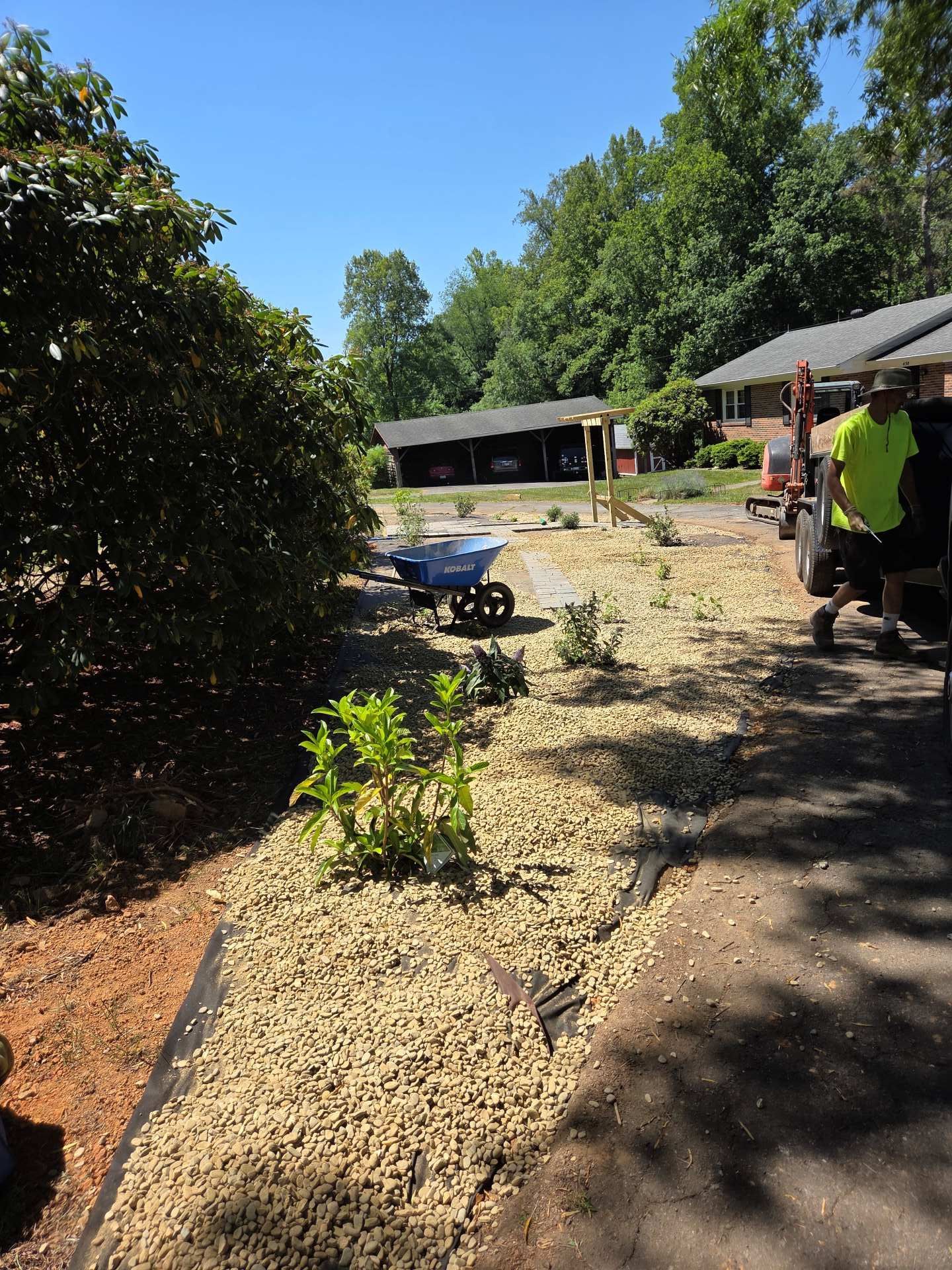 Sunny garden path beside a house, with wheelbarrow, shrubs, and a person in a neon shirt near the porch.