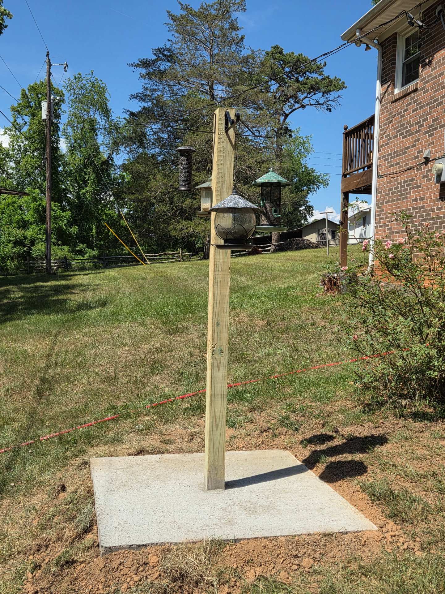Wooden post on a concrete pad in a grassy yard beside a brick house, with a hanging bird feeder.
