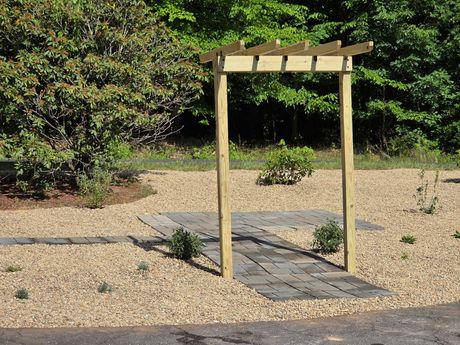 Wooden garden arbor over a stone path in a gravel landscape with green shrubs and trees.
