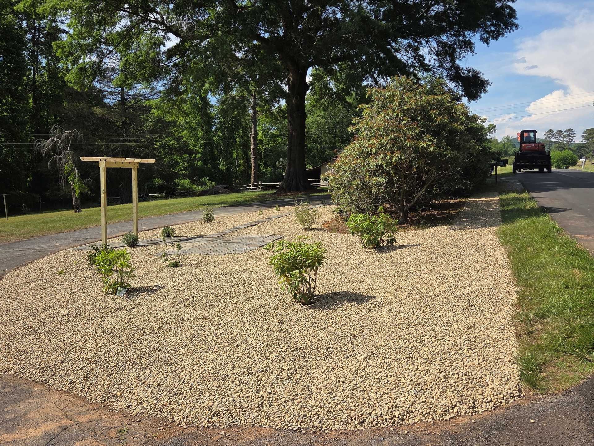 Gravel landscape bed with small shrubs and a wooden sign beside a road in bright sunlight