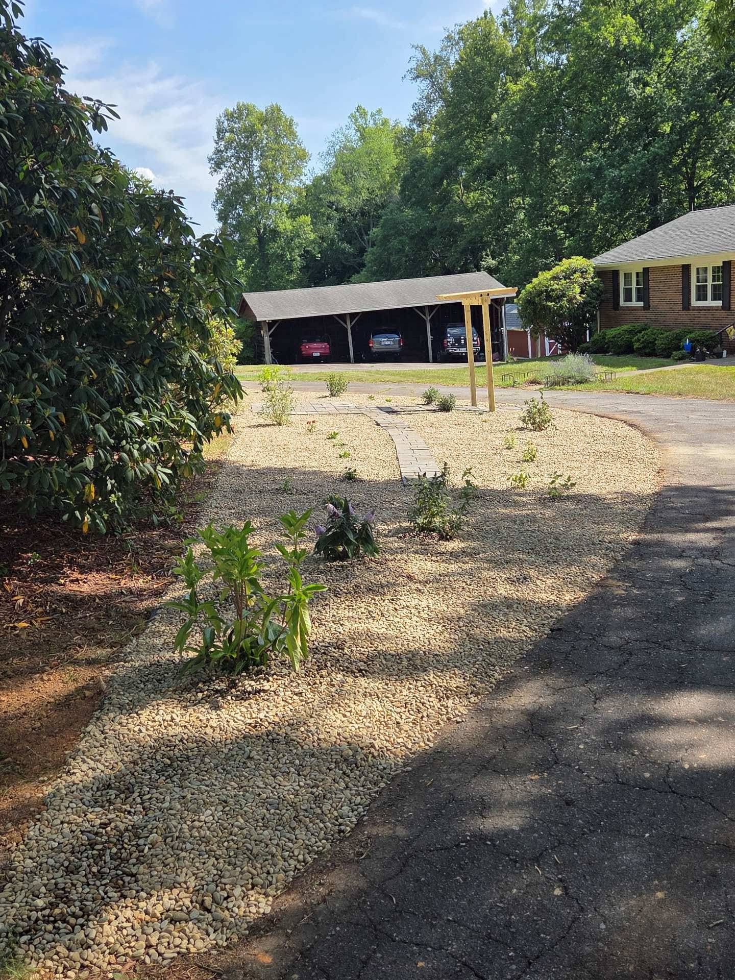 Gravel driveway leading to a carport and house, with trees and a sunlit front yard.