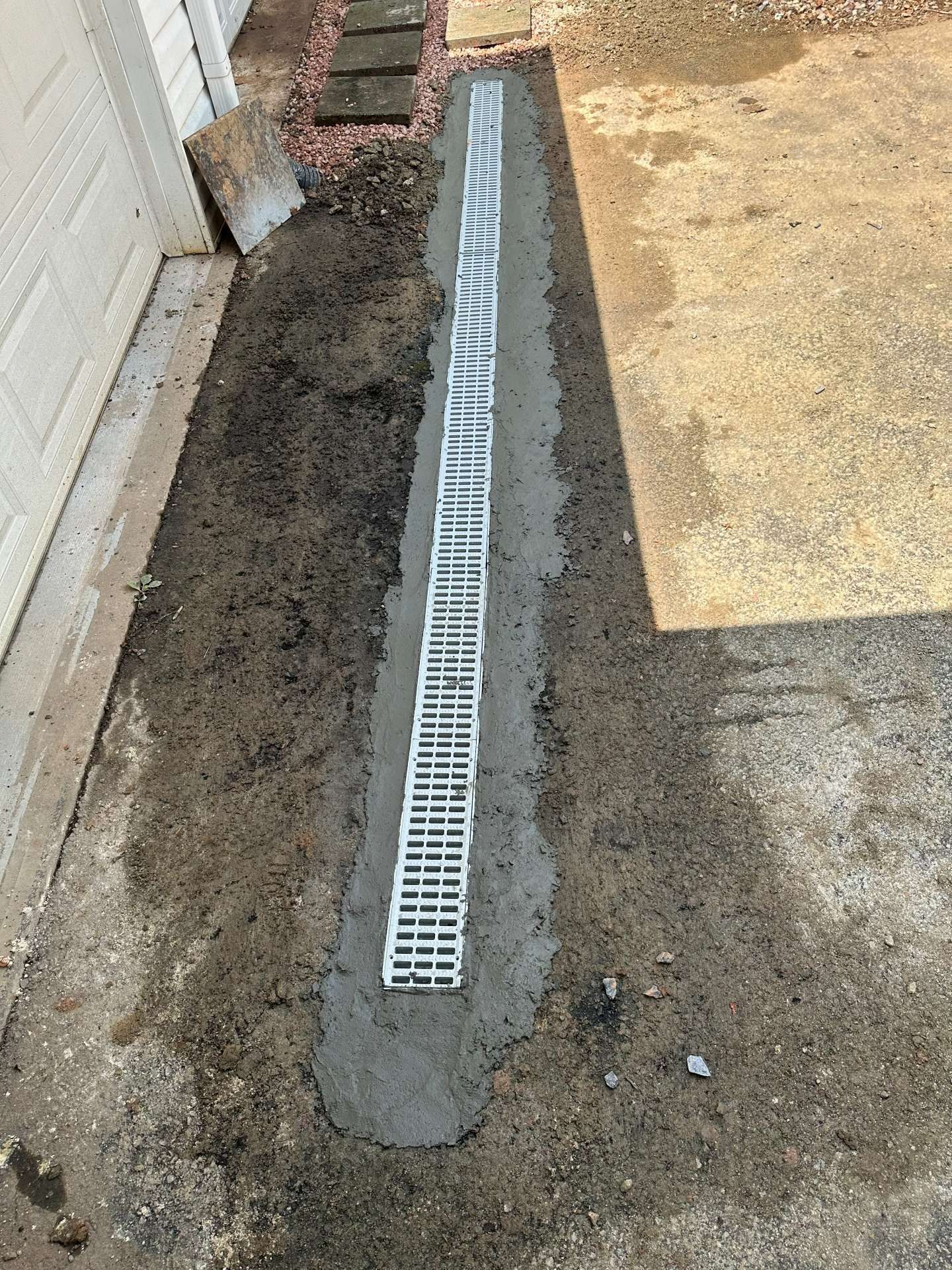 Narrow concrete drainage channel with a white grate between dirt and a paved walkway beside a wall