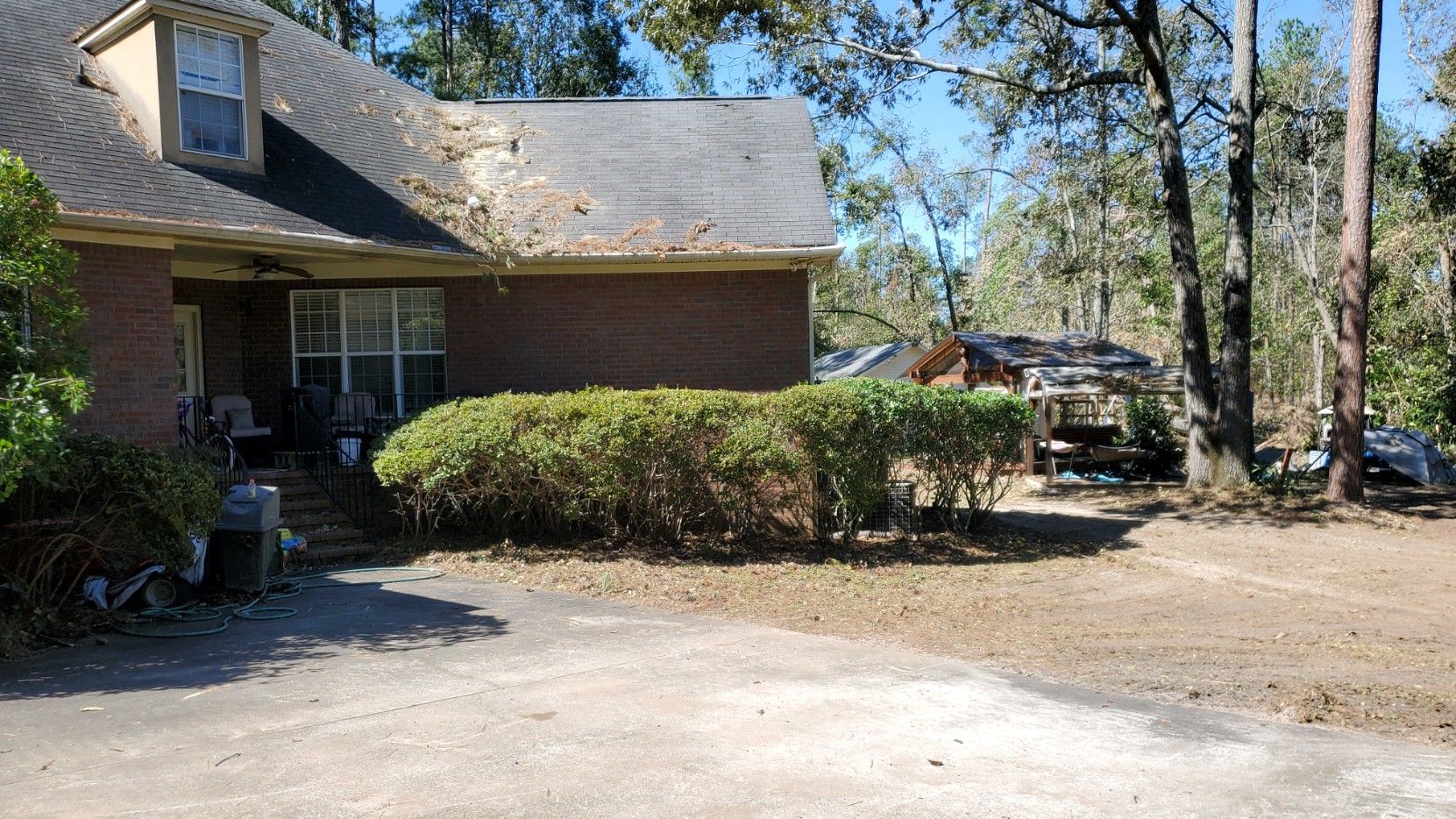 Brick house with a small front porch, trimmed hedges, and a gravel driveway in a wooded yard.