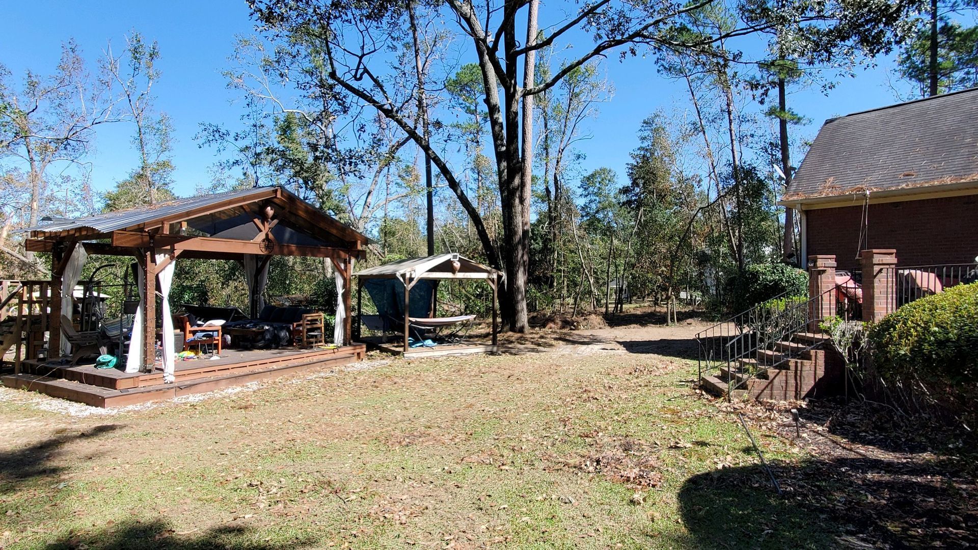 Backyard with a wooden gazebo, tall trees, and a brick house under a clear blue sky