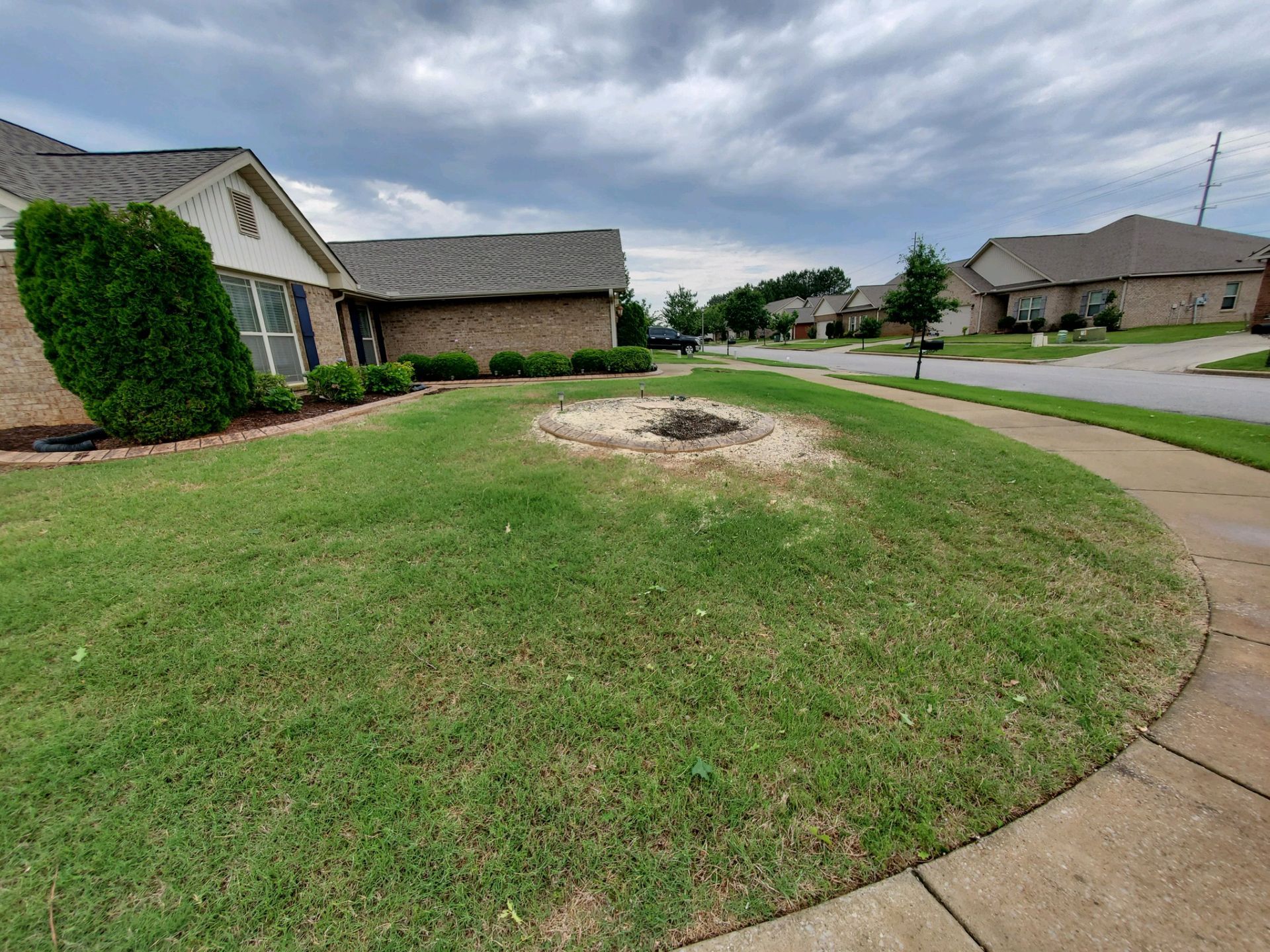 Suburban front yard with a large bare dirt patch in the center under cloudy skies.