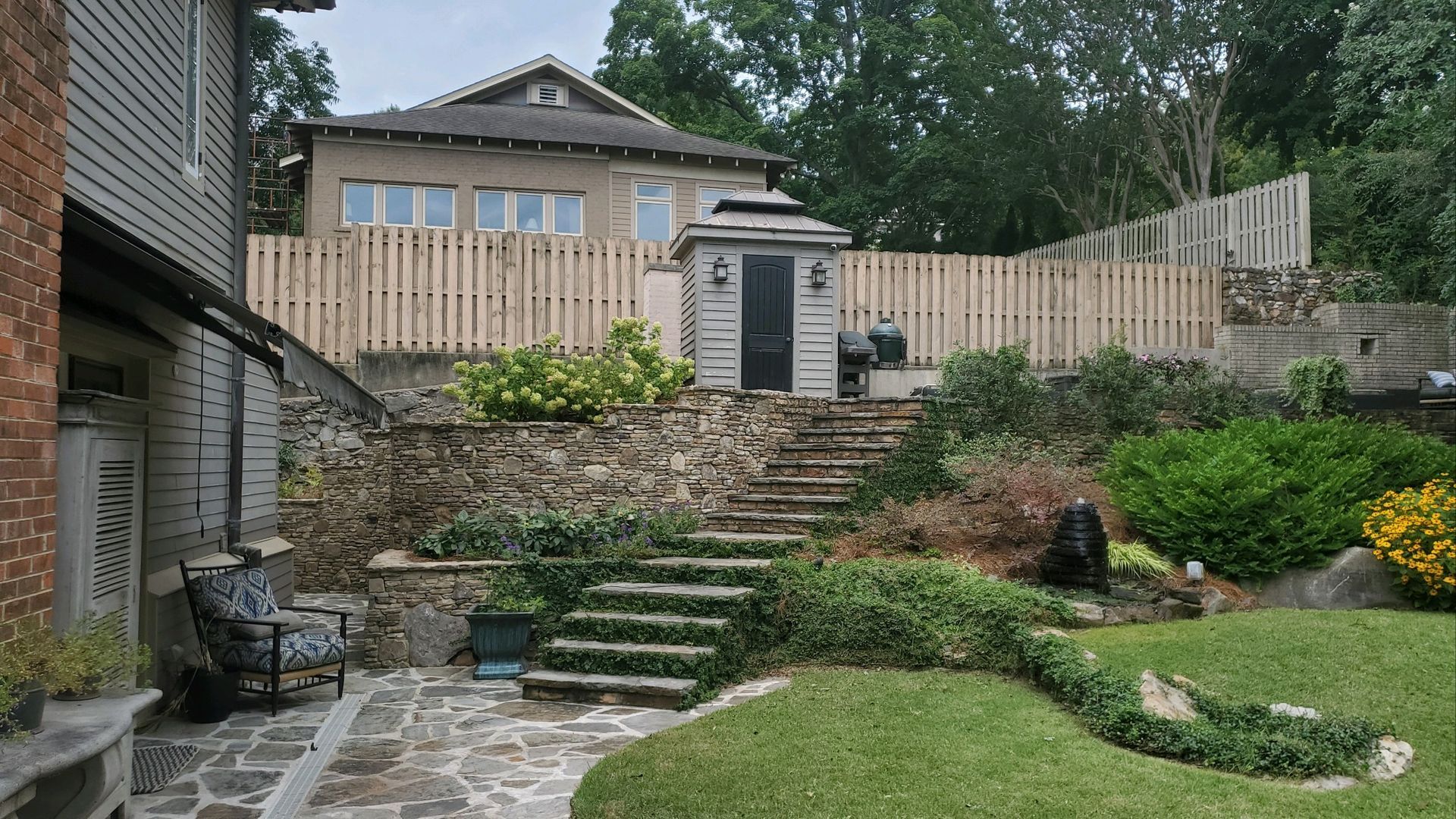 Backyard stone steps leading to a raised patio with wooden fence, lush garden, and house in the background