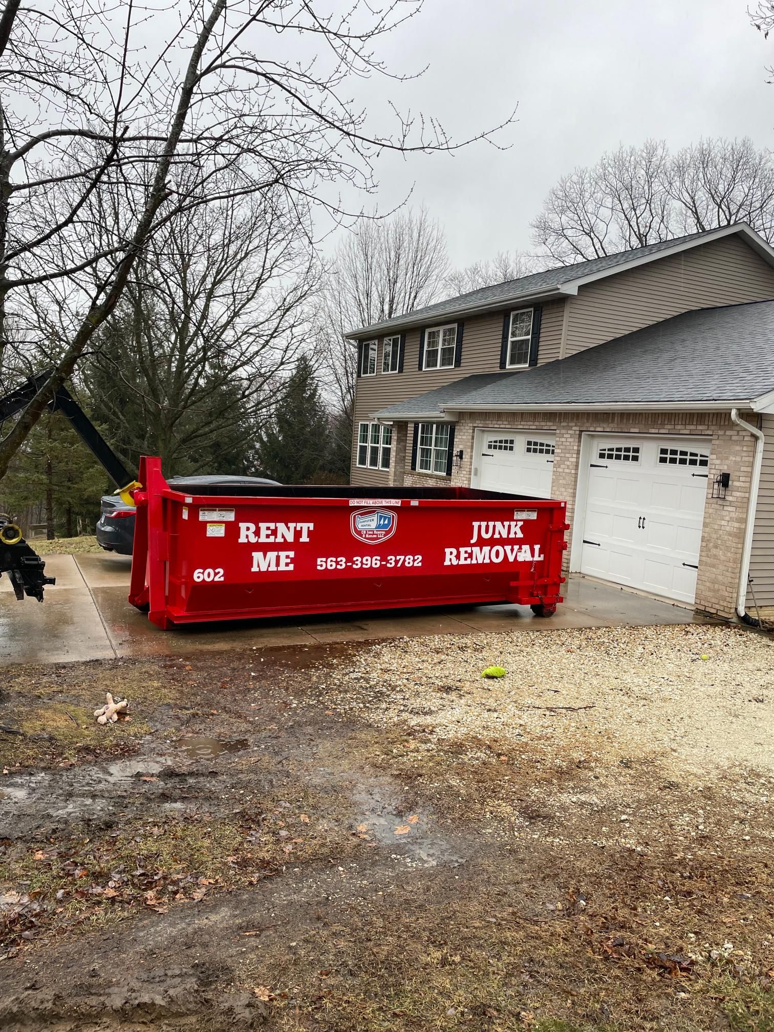 A bright red junk removal dumpster sits on a residential driveway in front of a house with a two-car garage.