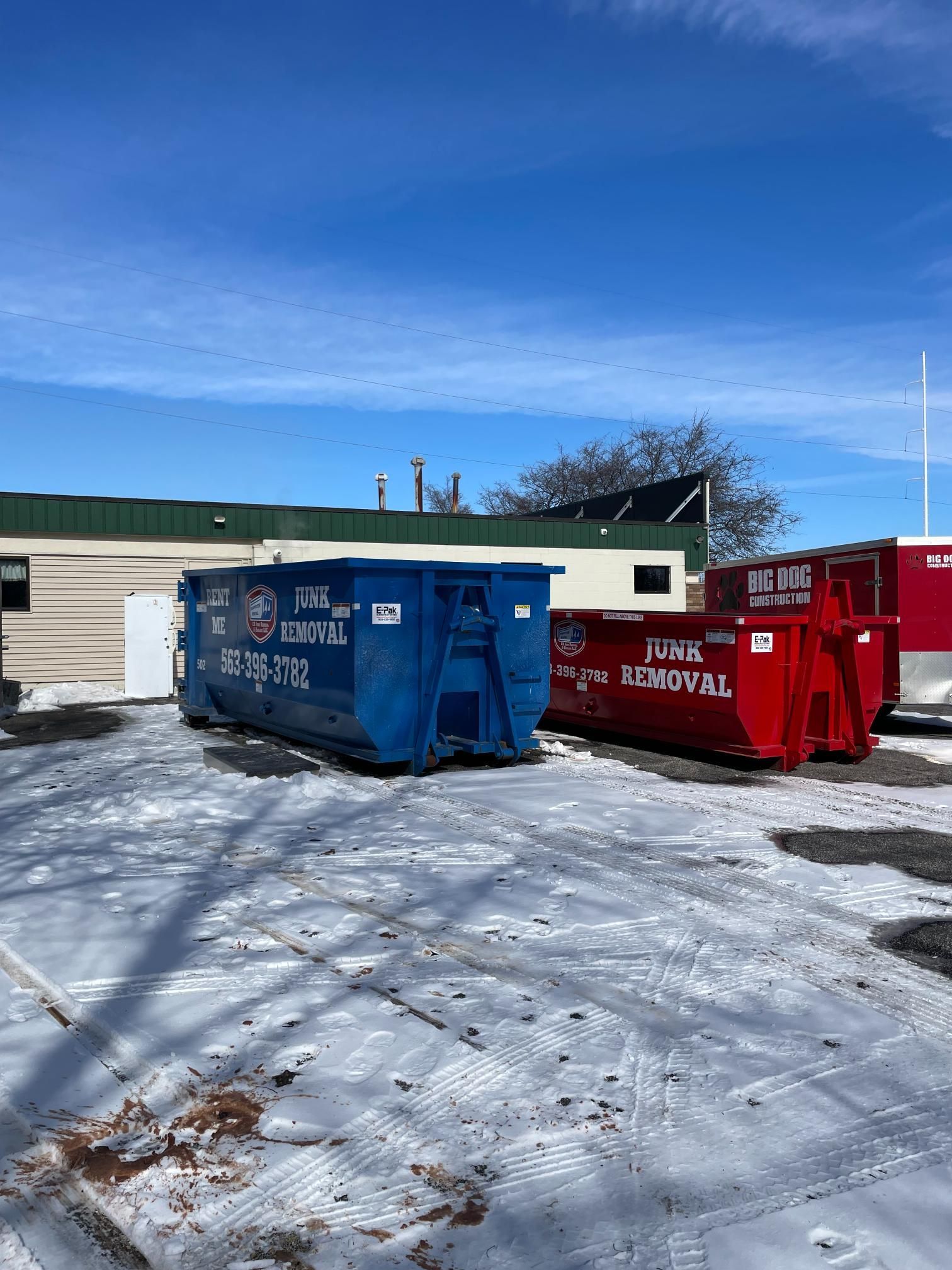 Blue and red dumpsters for junk removal parked on a snowy lot in front of a building under a bright blue sky.