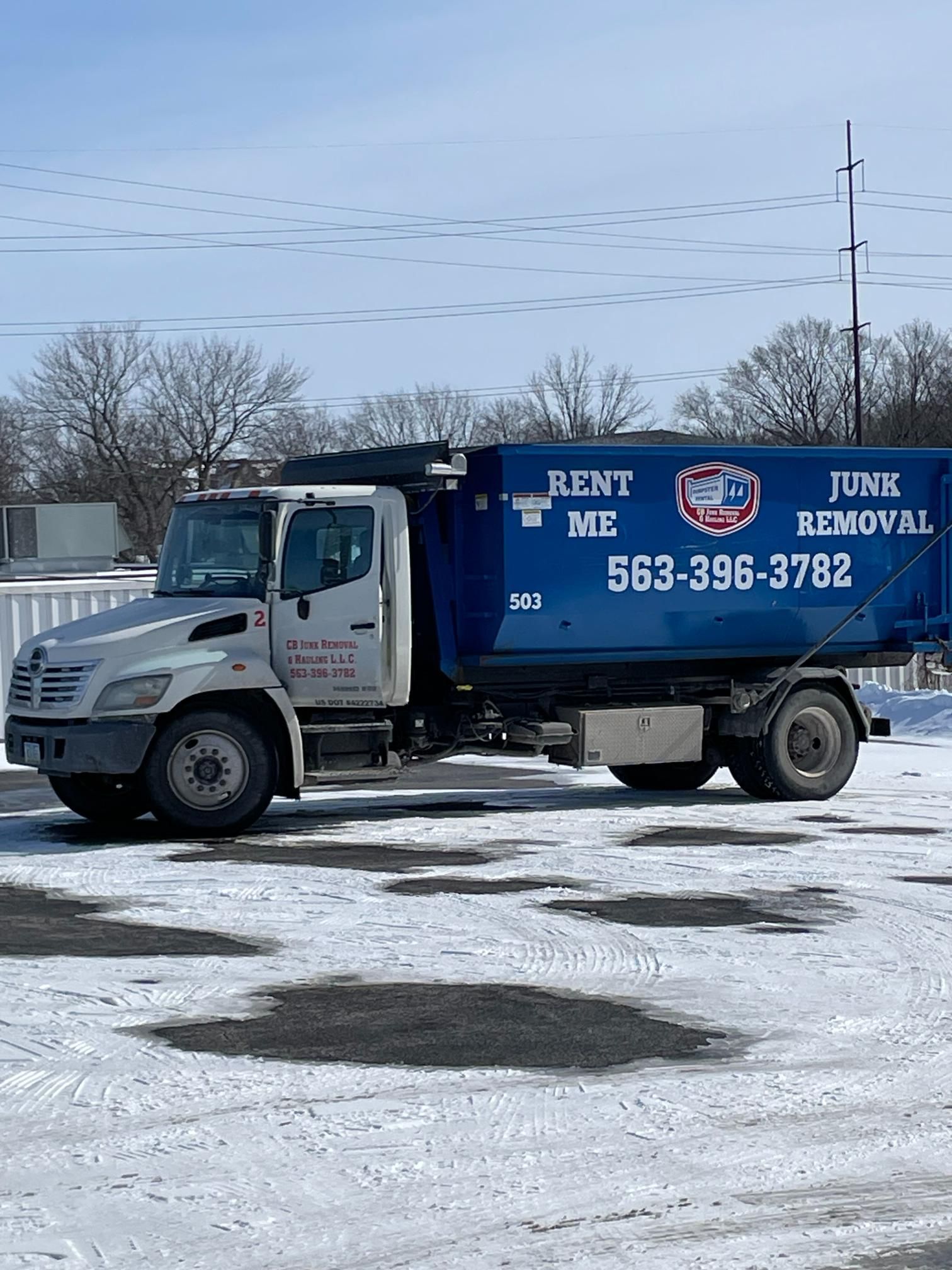 A white junk removal truck parked on a snowy, open lot, featuring a large blue dumpster labeled with a phone number.