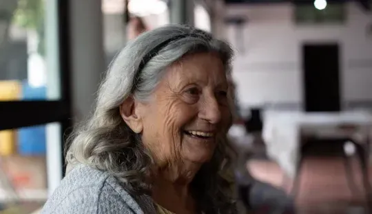 An elderly woman with gray hair is smiling while sitting in front of a window.