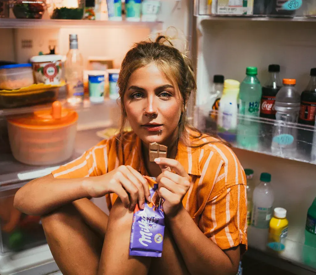 A woman is sitting in front of an open refrigerator eating a milka bar.