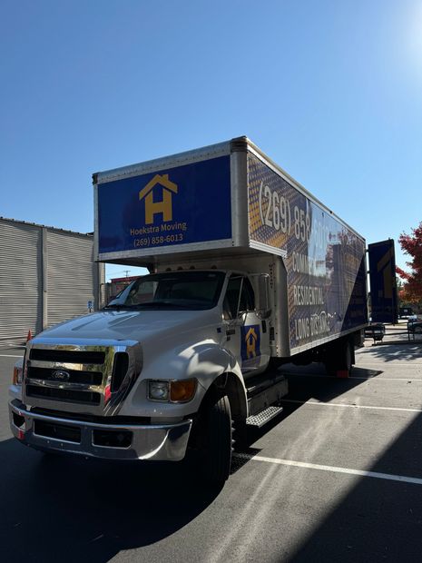 White moving truck parked outdoors on a sunny street, with a blue cargo box and house logo.