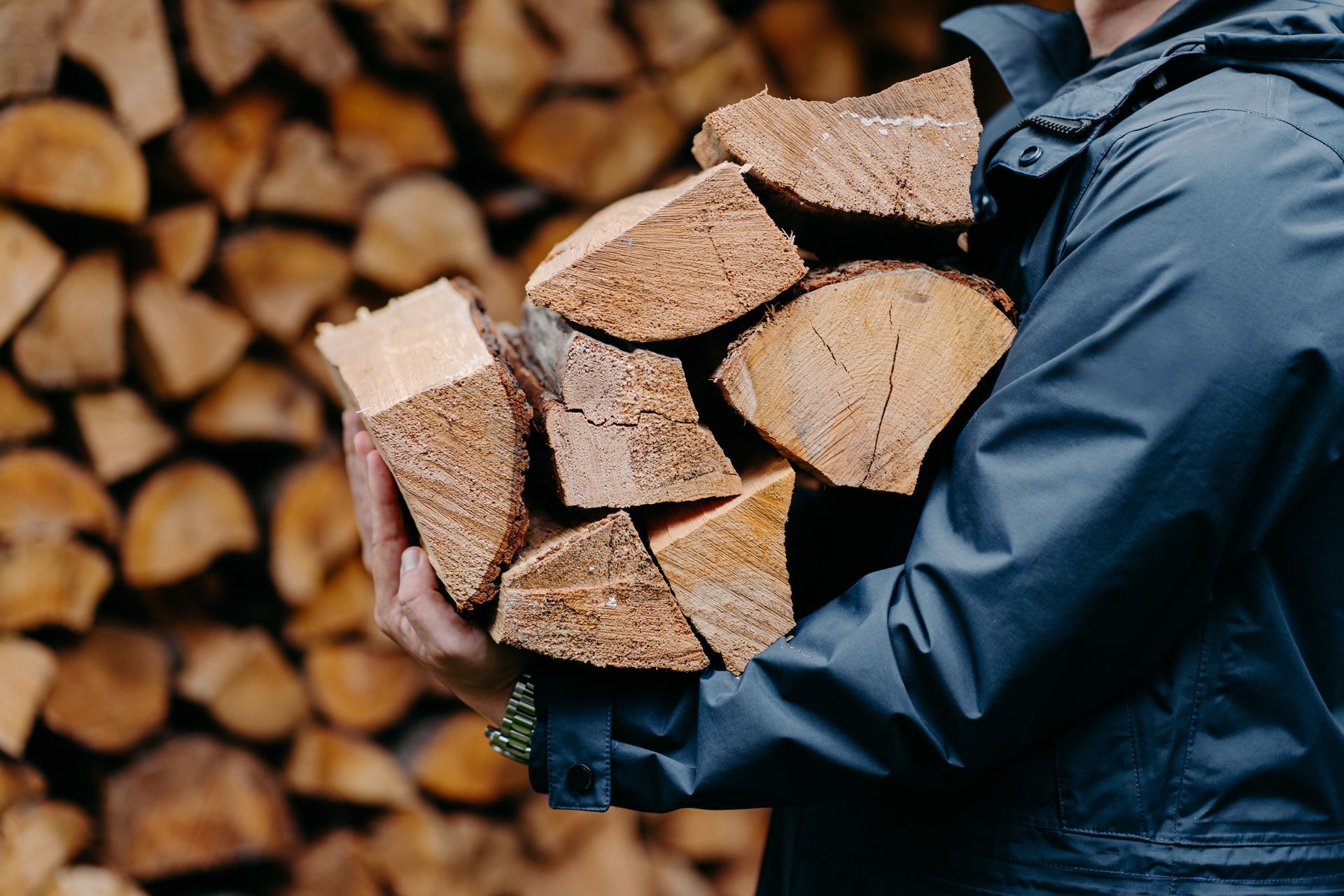 Person carrying split firewood in a dark jacket, with stacked logs blurred in the background