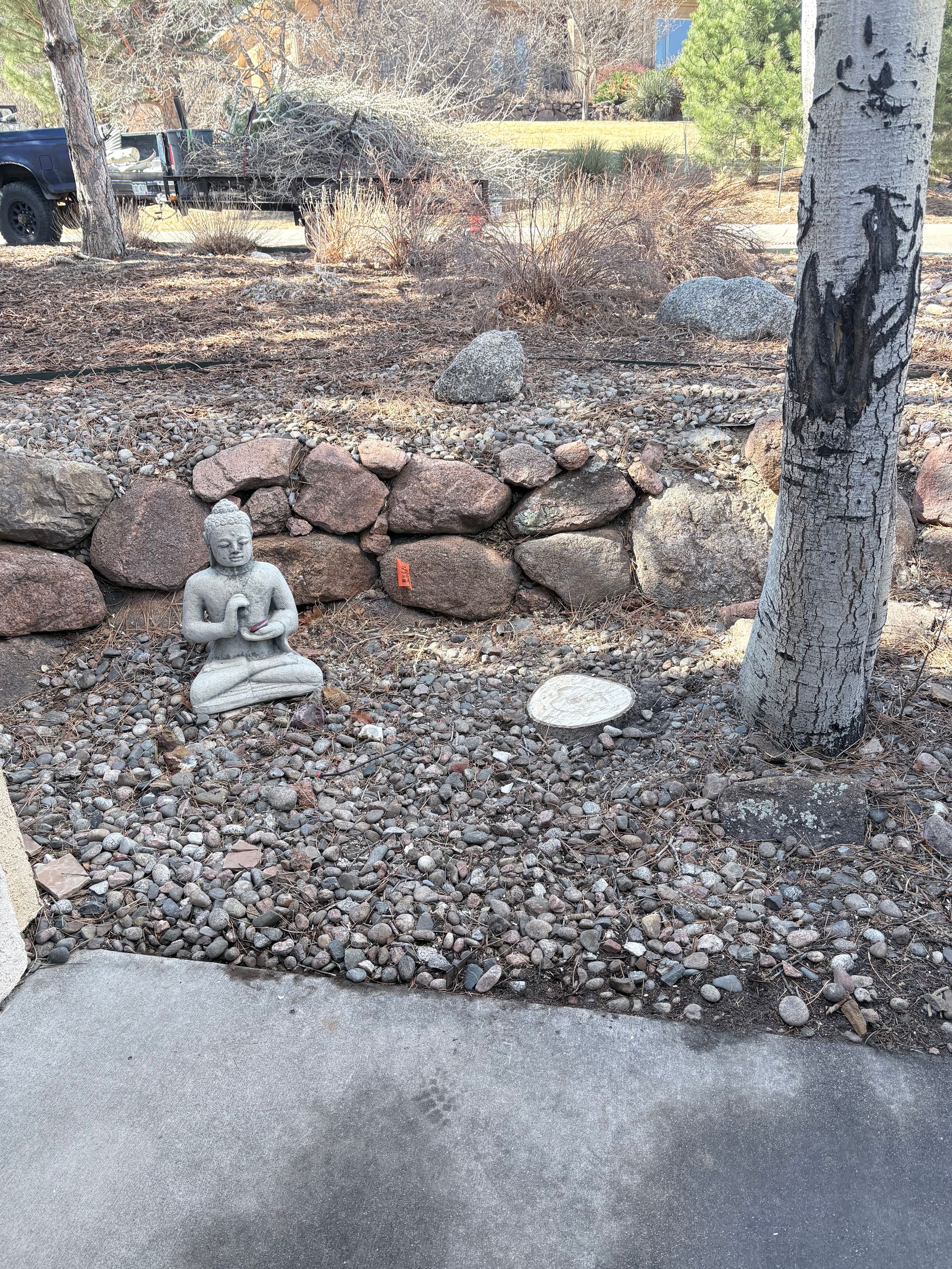 Small Buddha statue by a rock wall in a gravel yard beside a tree and concrete path