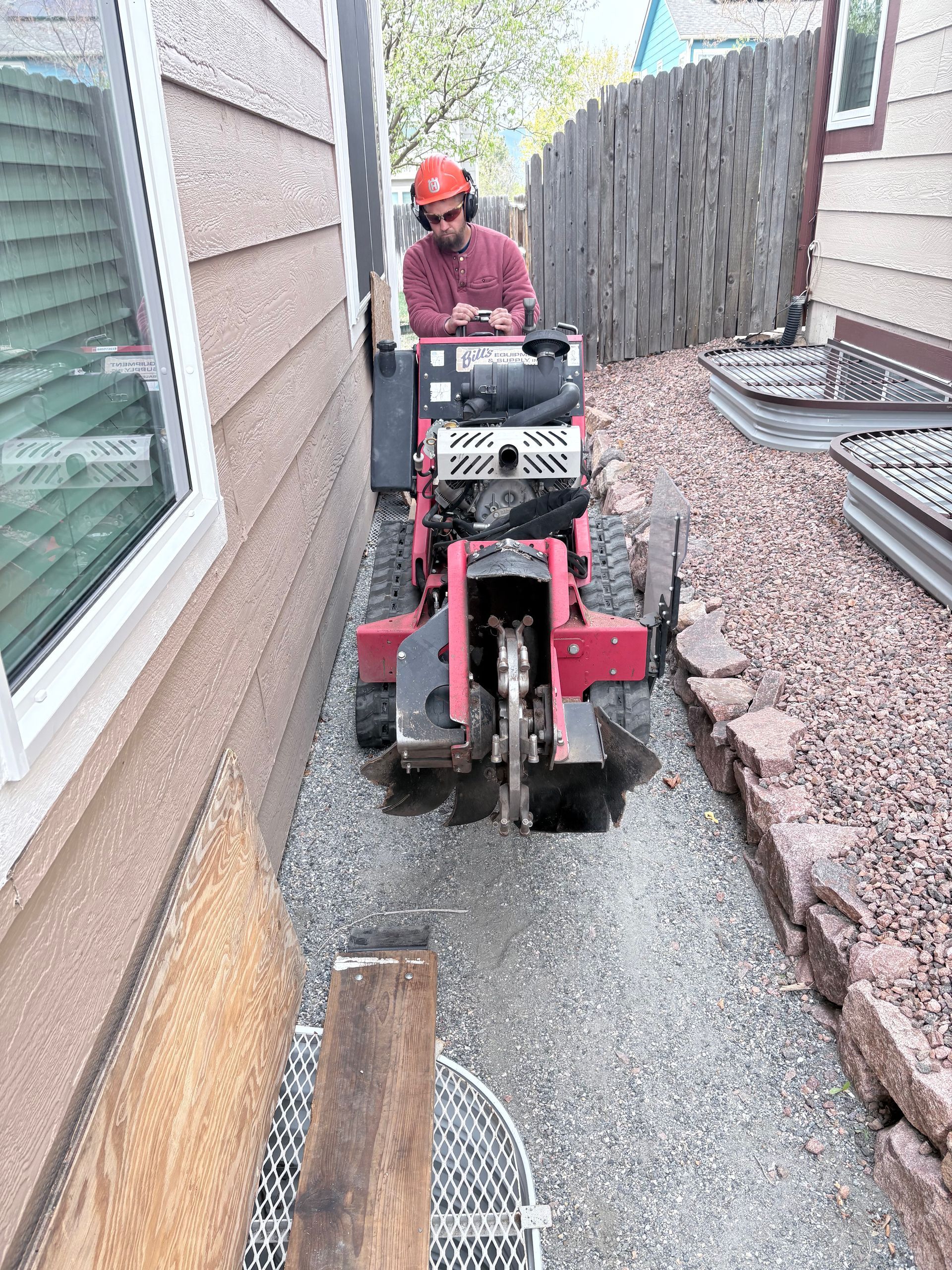 Worker operating a red mini excavator in a narrow gravel side yard beside a house