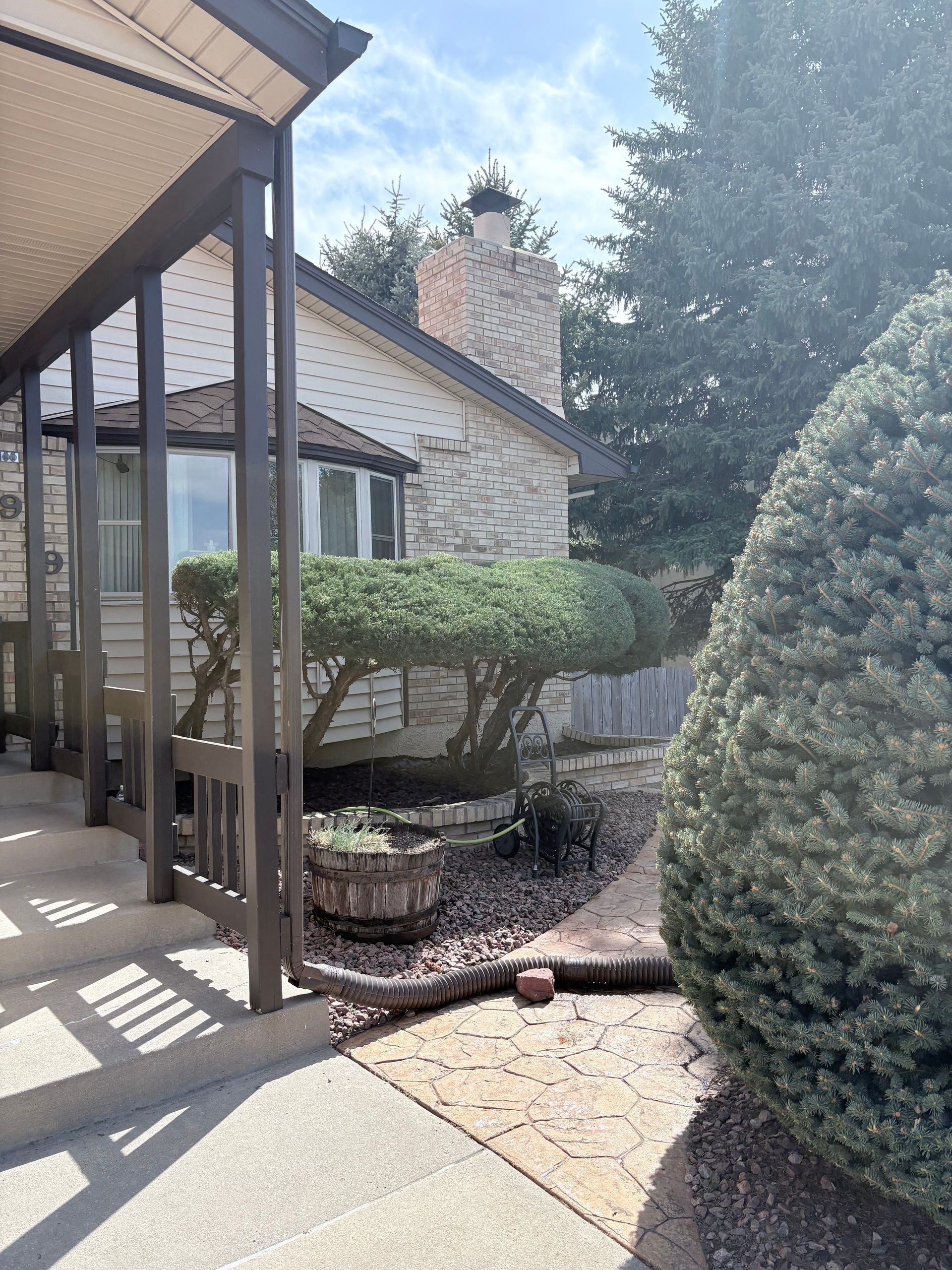 Covered patio beside a brick house, with trimmed shrubs, a small planter, and a gravel garden path.