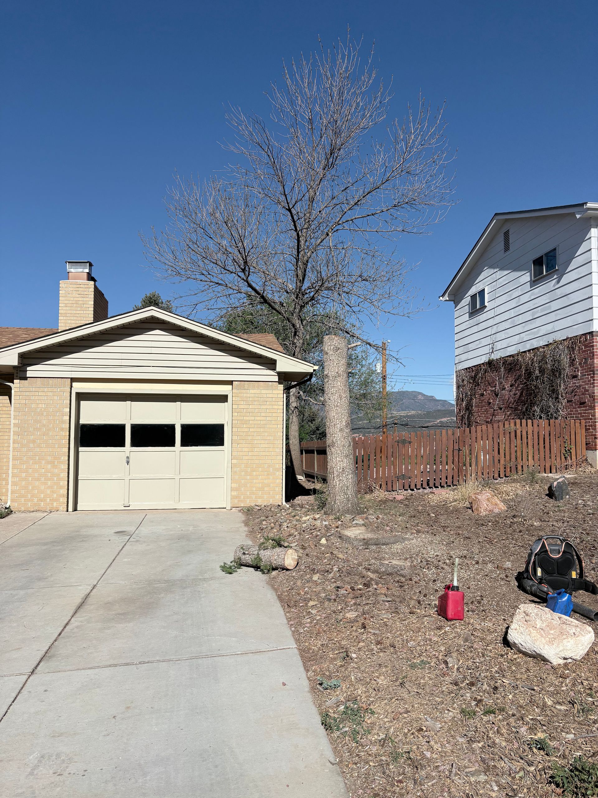 Suburban driveway beside a garage and house, with leafless tree, fence, and small yard items.