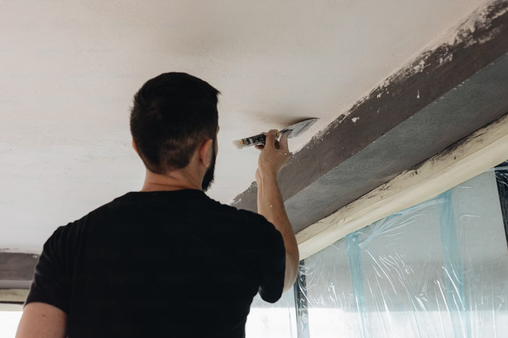 Man using a scraper on a ceiling, near a dark beam and window, indoors.