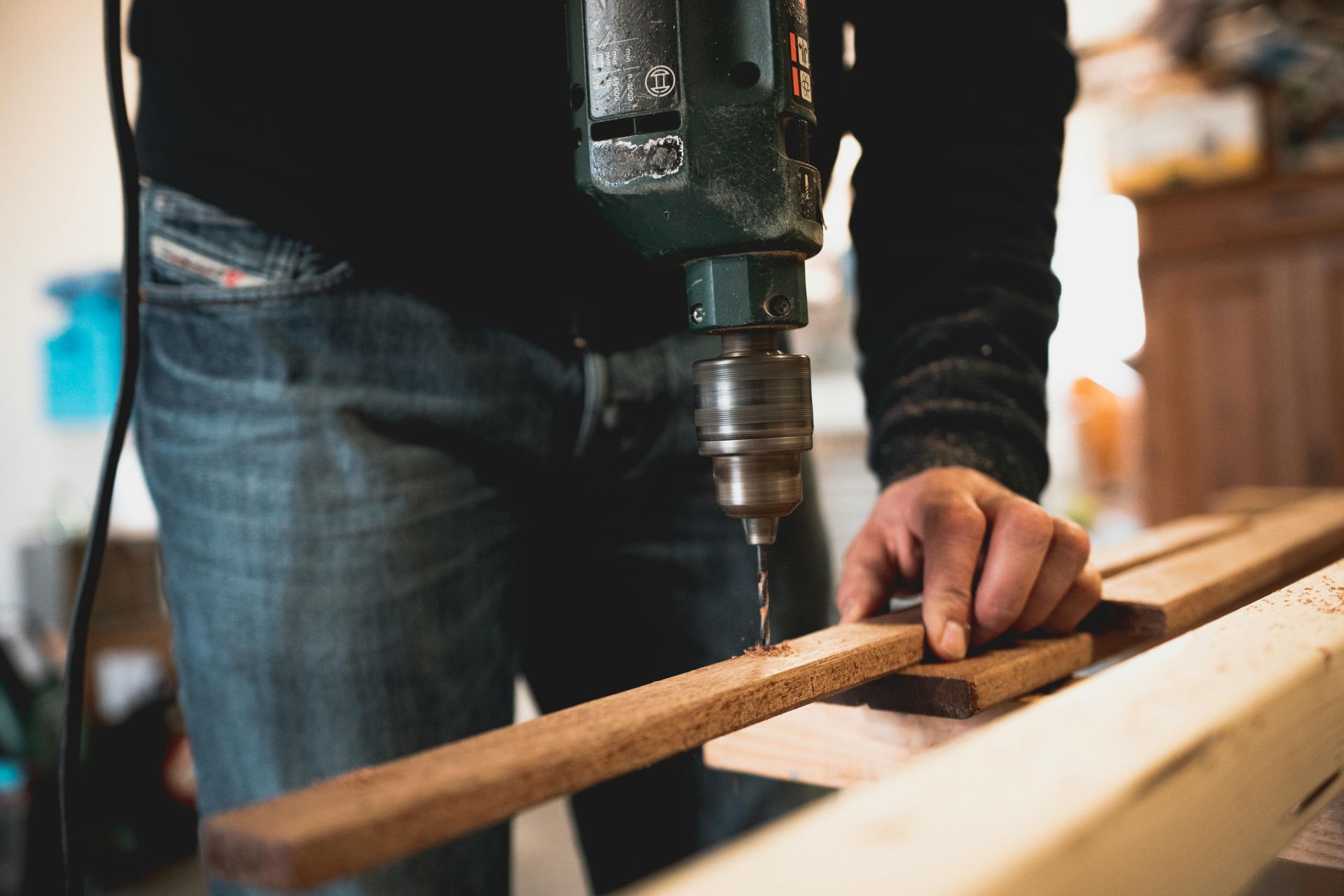 A person uses a power drill to bore a hole into a thin strip of wood in a workshop setting.