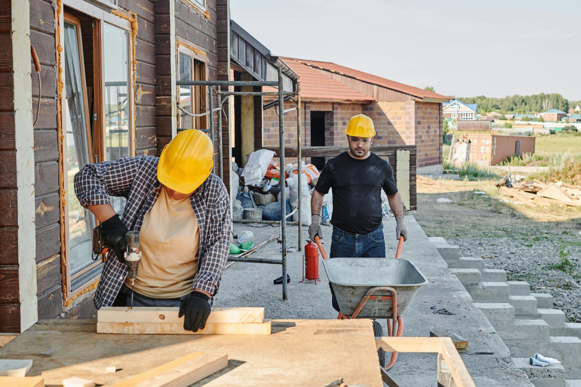 Two workers in yellow hard hats at a construction site; one saws a wooden board while the other pushes a wheelbarrow.
