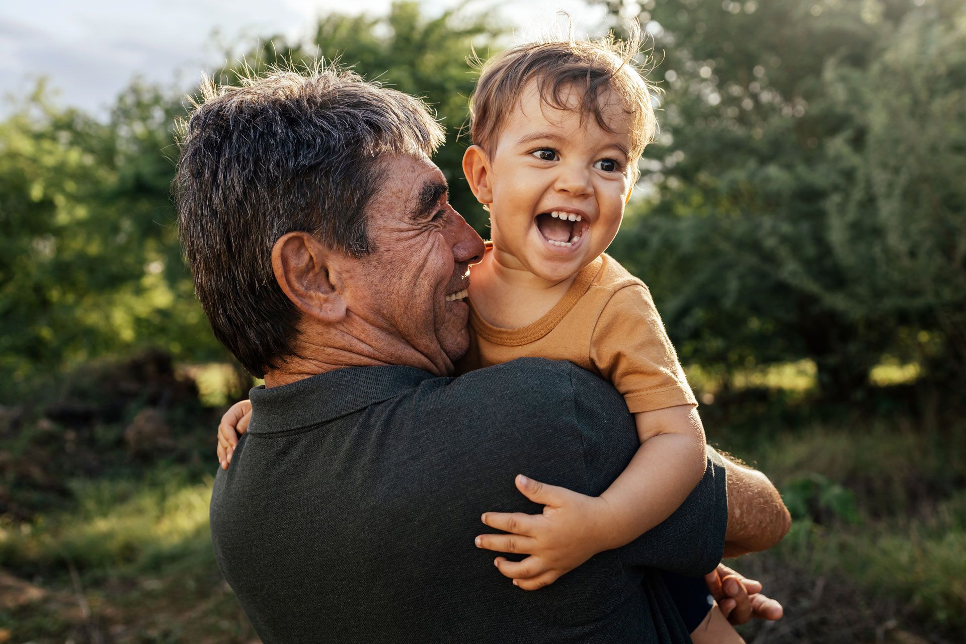 Man holding and smiling at a laughing toddler outdoors.