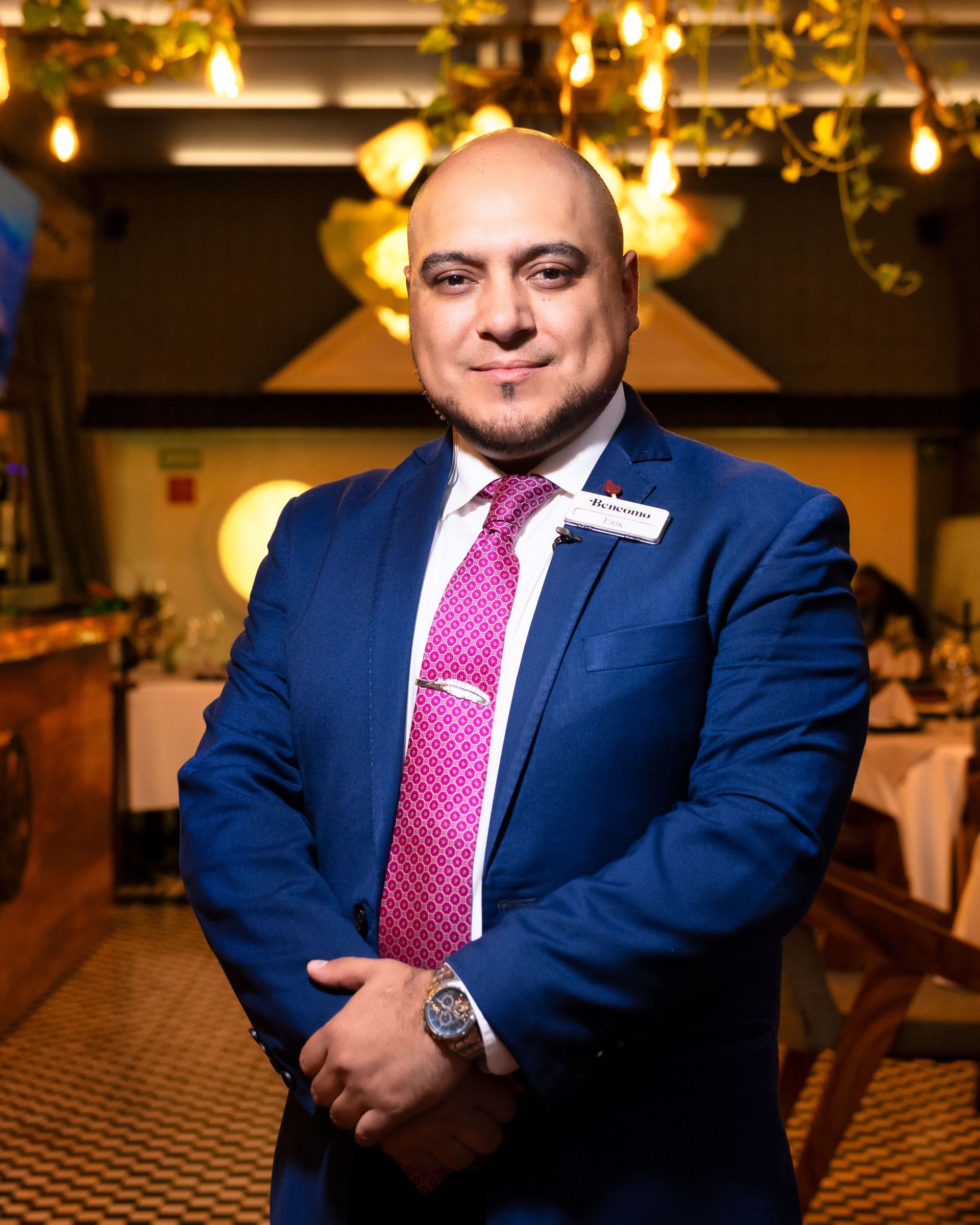 Man in a blue suit and pink tie standing indoors with warm hanging lights in the background