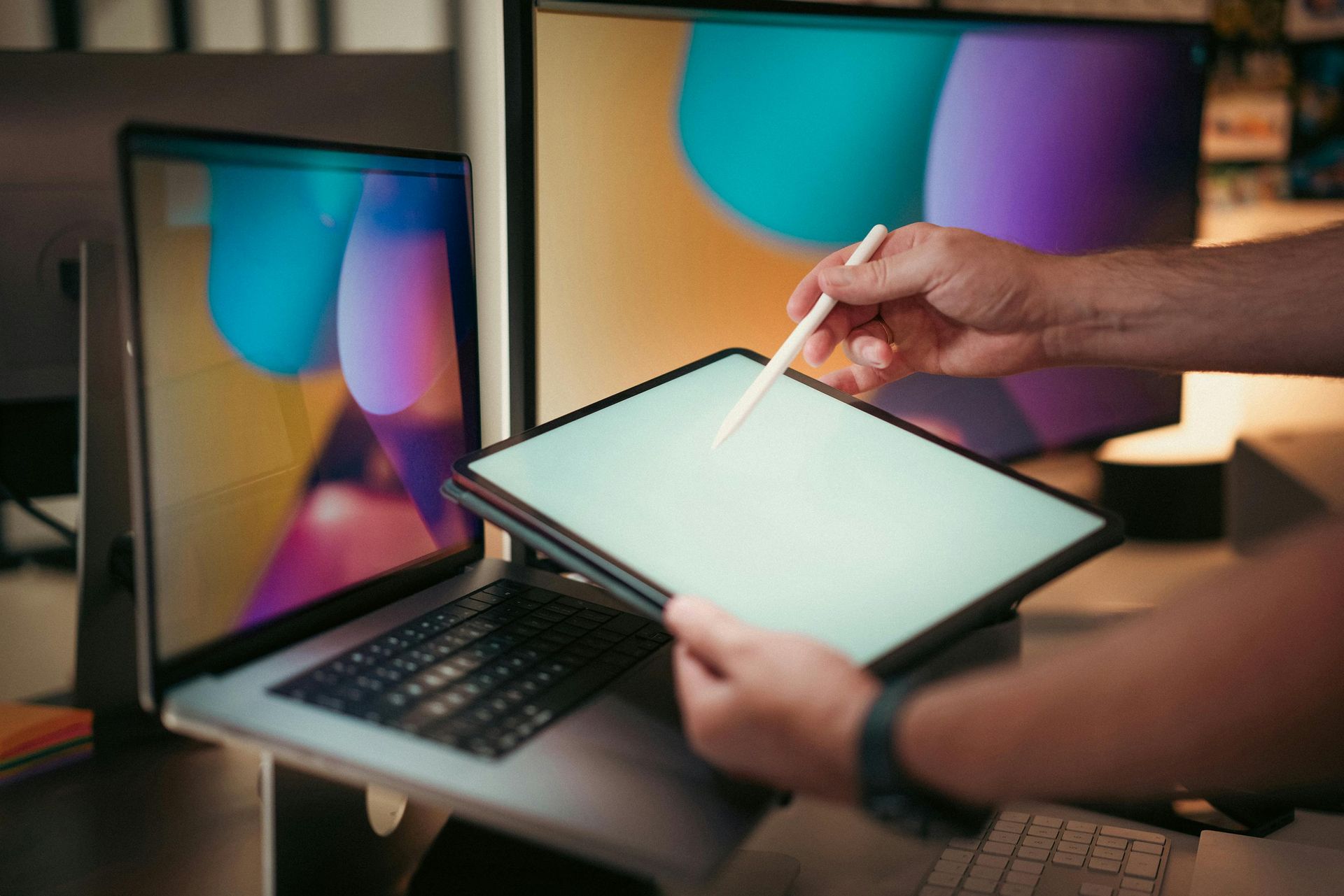 Person using a stylus on a tablet beside a laptop displaying colorful abstract graphics
